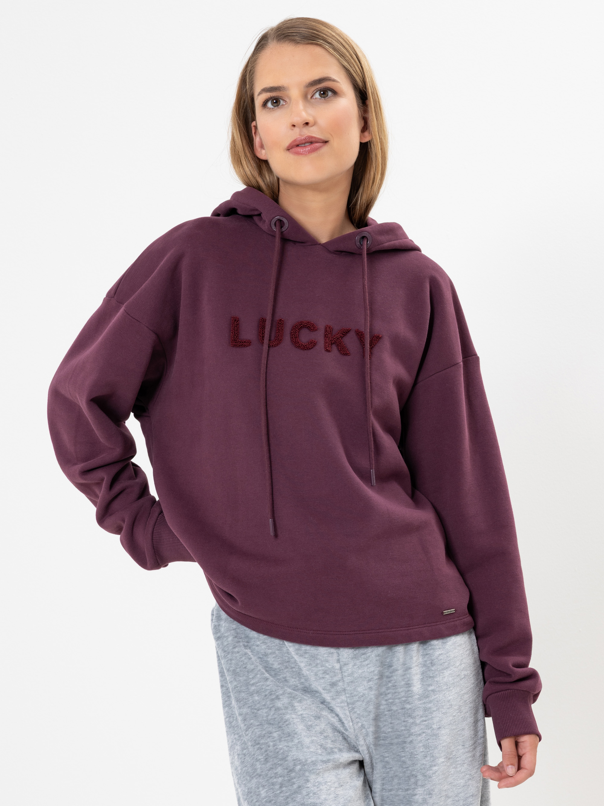 Eine Frau mit blonden Haaren trägt einen kastanienbraunen Kapuzenpullover mit der Aufschrift LUCKY auf der Vorderseite und eine hellgraue Jogginghose vor einem schlichten weißen Hintergrund.