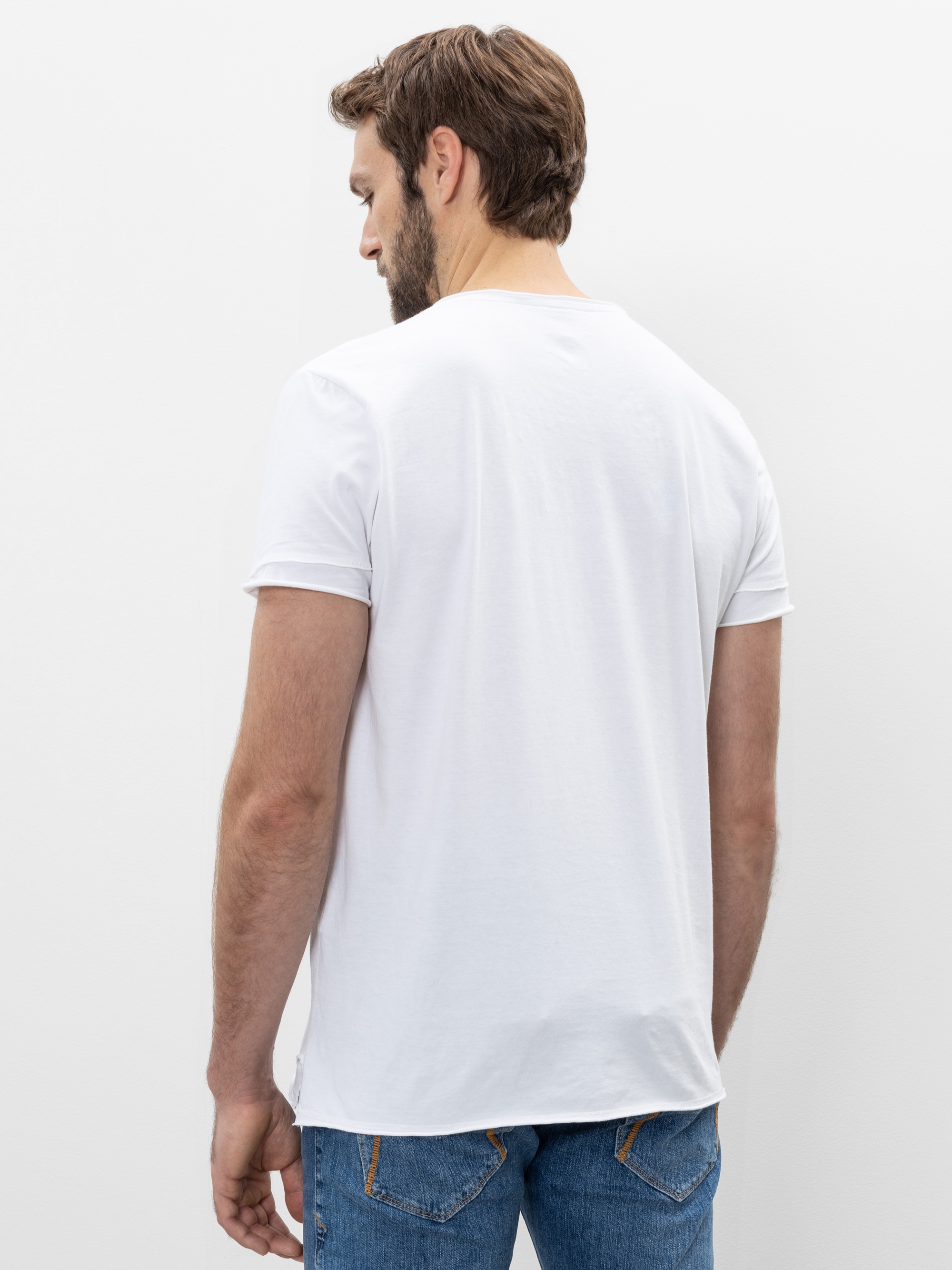 Ein Mann mit braunem Haar und Bart steht mit dem Rücken zur Kamera, trägt ein schlichtes weißes T-Shirt und blaue Jeans vor einem weißen Hintergrund.