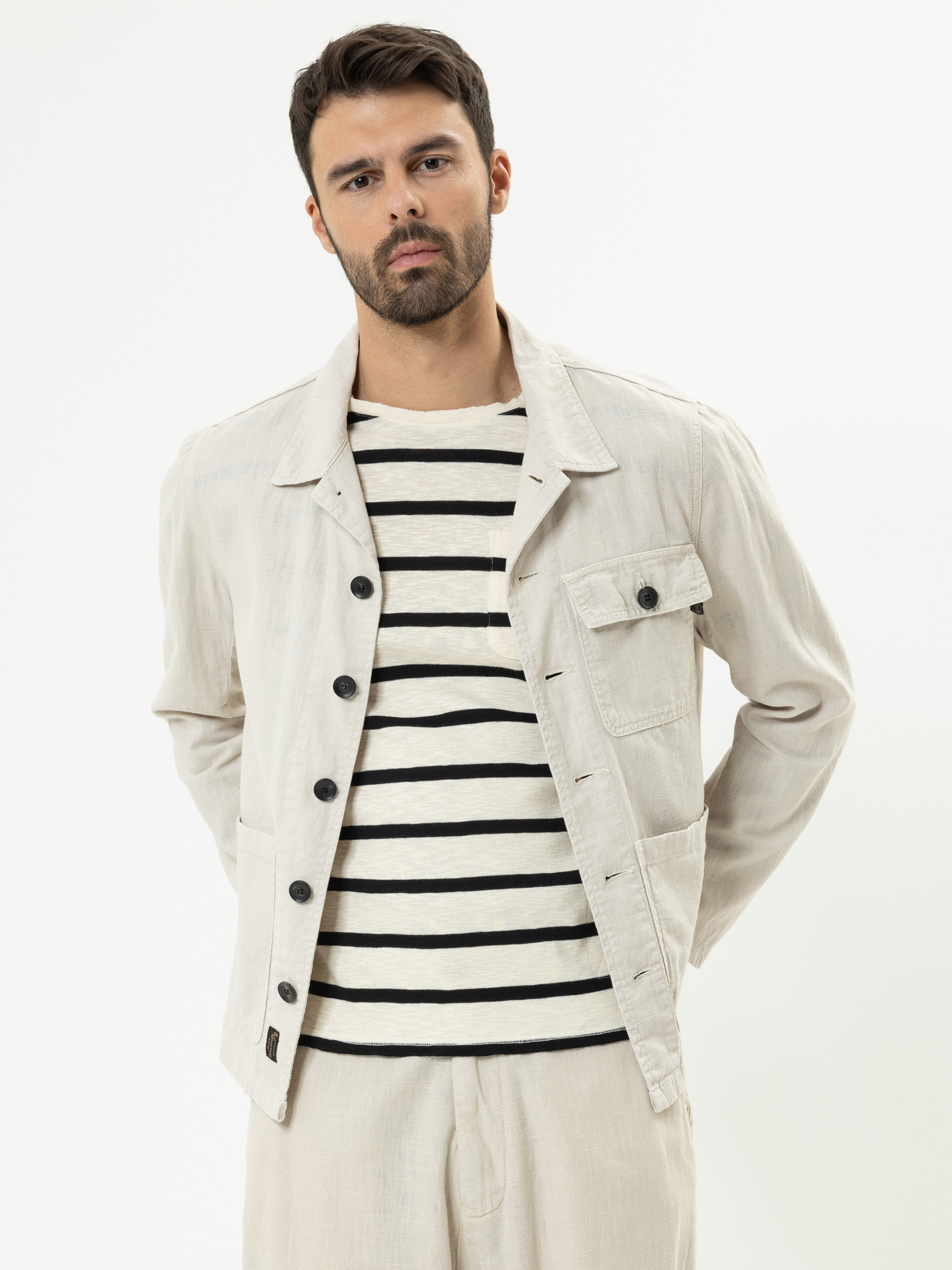 Ein Mann mit dunklen Haaren und einem Bart trägt eine hellbeige Jacke über einem cremefarbenen und schwarz gestreiften Hemd, gepaart mit einer hellbeigen Hose. Er steht vor einem schlichten weißen Hintergrund und blickt mit neutraler Miene in die Kamer