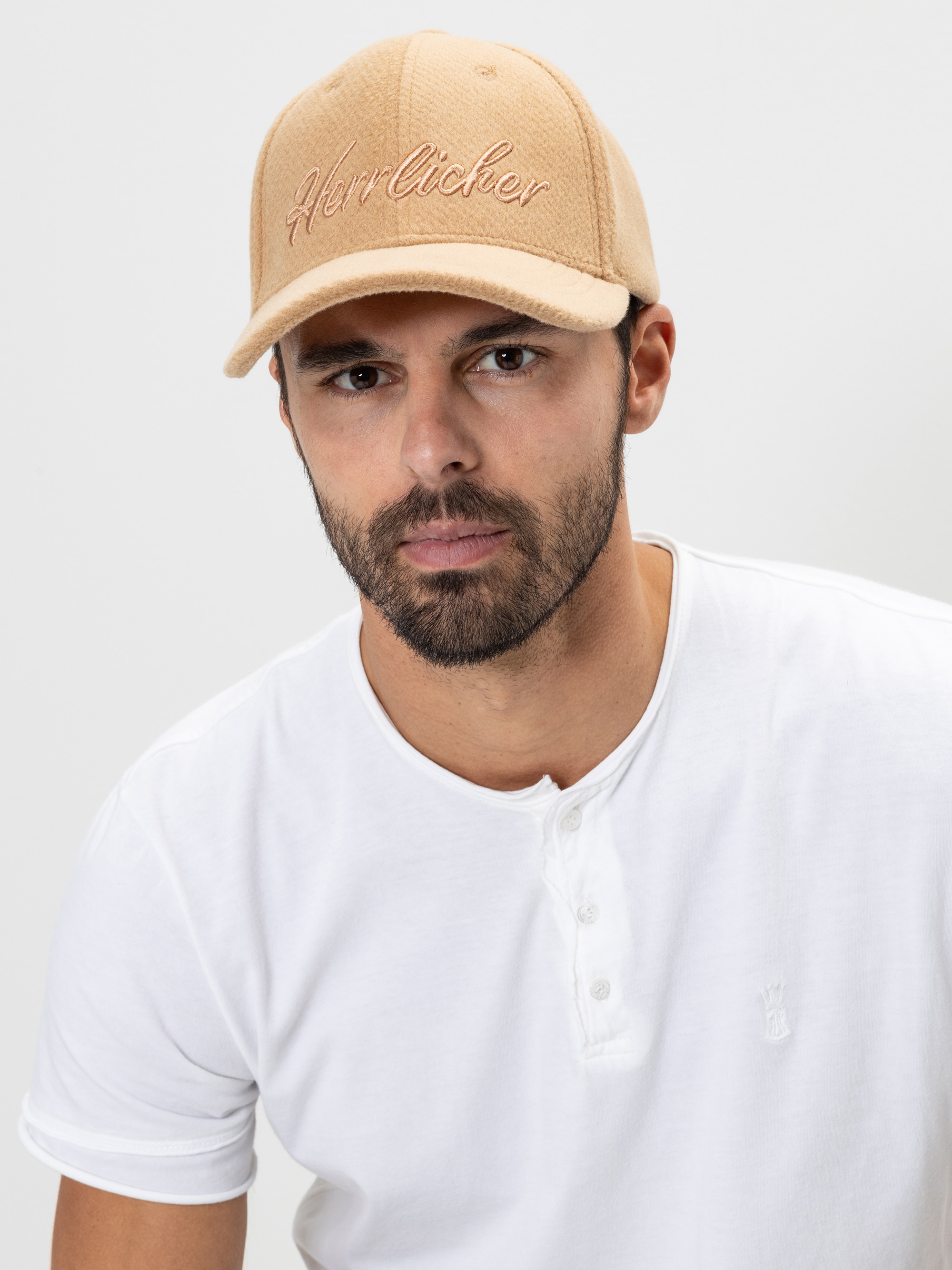 Ein Mann mit Bart, der eine beigefarbene Mütze mit der Aufschrift "Herrlicher" und ein weißes Hemd trägt und vor einem schlichten hellen Hintergrund direkt in die Kamera blickt.