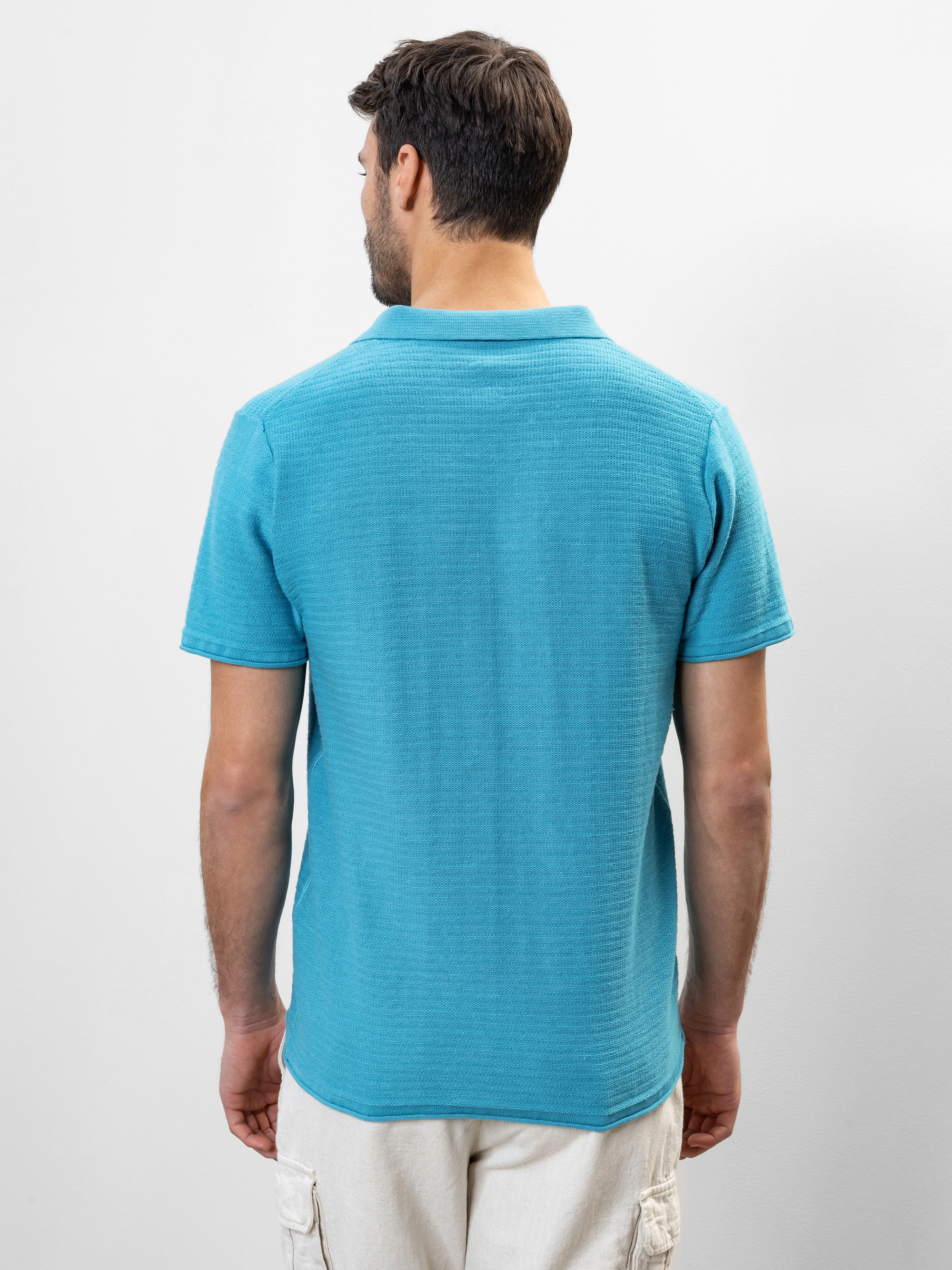 Ein Mann mit kurzen braunen Haaren, bekleidet mit einem hellblauen kurzärmeligen Polohemd und einer cremefarbenen Hose, steht vor einem einfarbigen hellen Hintergrund und blickt von der Kamera weg.