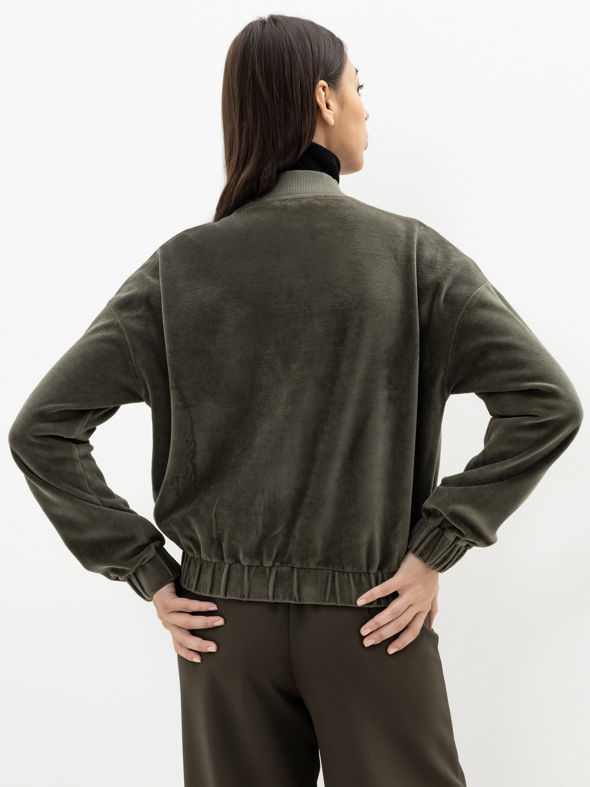 Eine Frau steht mit dem Rücken zur Kamera in einer olivgrünen, langärmeligen, samtigen Jacke und einer passenden Hose, die Hände in die Hüften gestemmt, vor einem schlichten weißen Hintergrund.