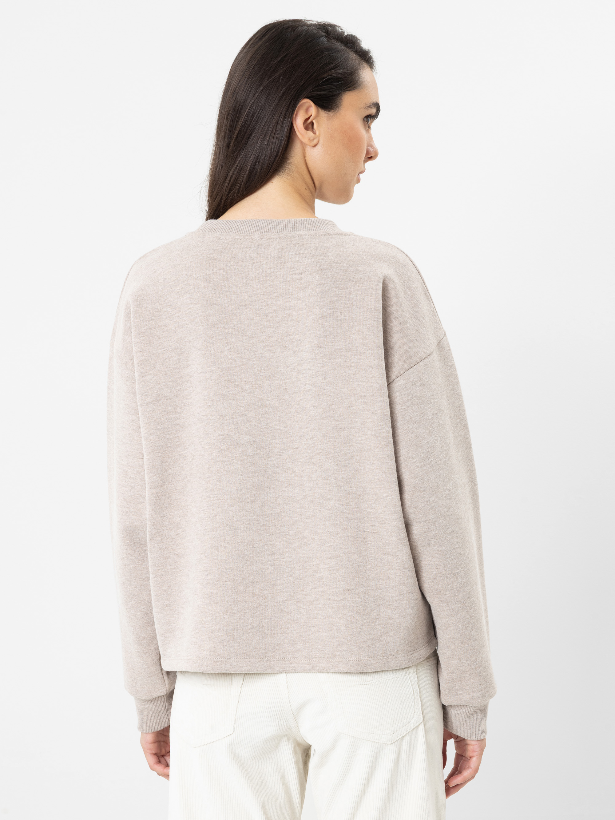 Eine Frau mit langen, dunklen Haaren, die einen lockeren, hellbeigen Pullover und eine weiße Hose trägt, steht mit dem Rücken zur Kamera vor einem schlichten weißen Hintergrund.
