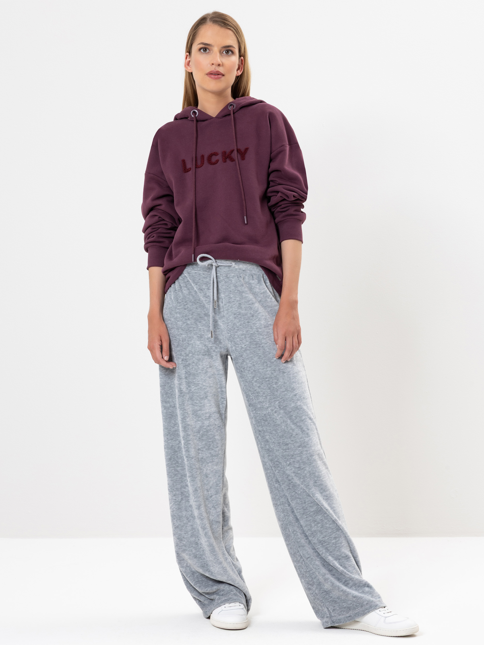 Eine Frau steht vor einem weißen Hintergrund und trägt einen kastanienbraunen Kapuzenpullover mit dem Aufdruck LUCKY, eine lockere hellgraue Jogginghose und weiße Turnschuhe. Sie hat glattes, hellbraunes Haar und einen entspannten Gesichtsausdruck.