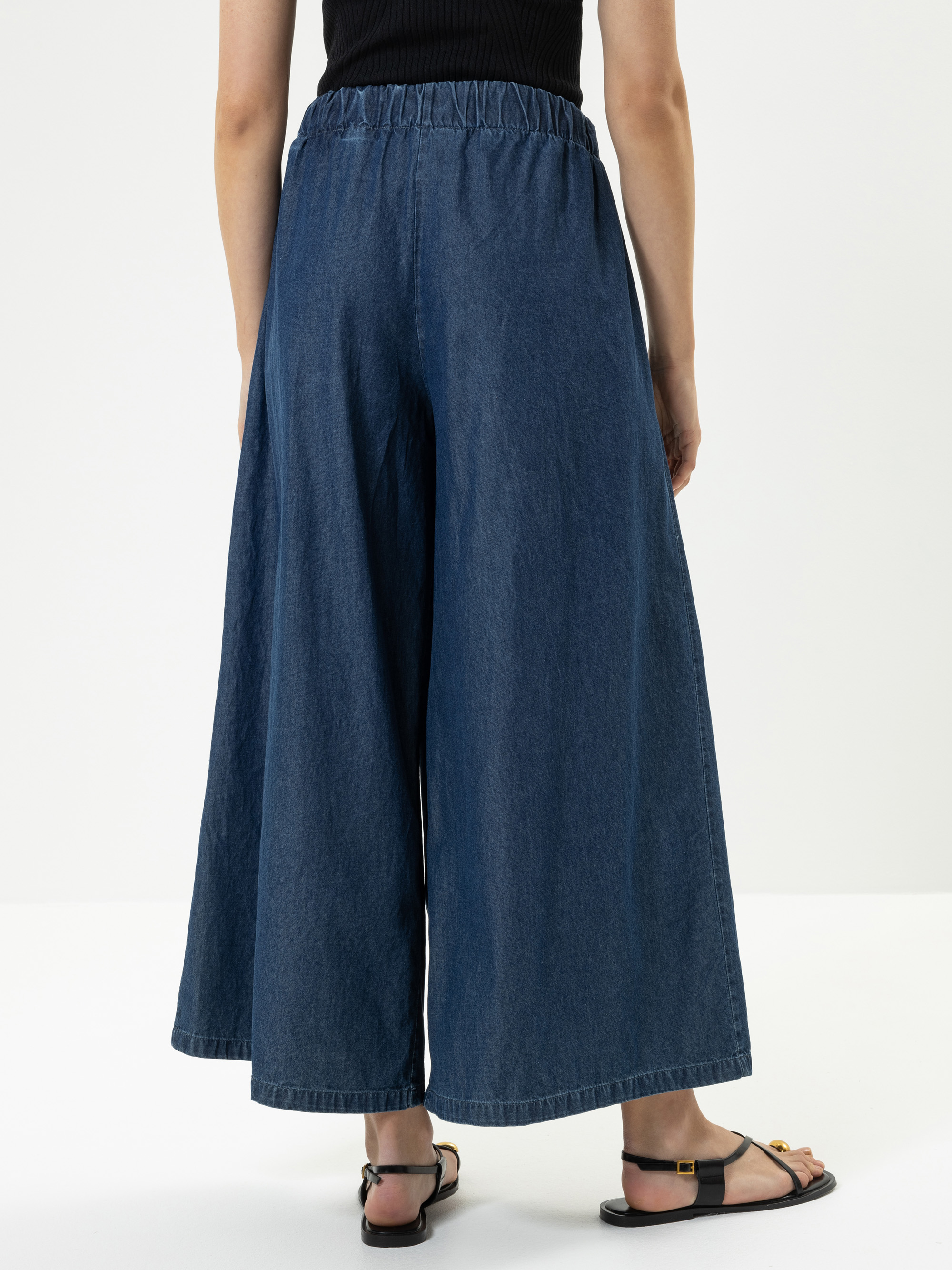 Eine Person, die eine knöchellange Jeanshose mit weitem Bein und elastischem Bund, ein schwarzes ärmelloses Oberteil und schwarze Sandalen trägt, steht vor einem schlichten weißen Hintergrund.