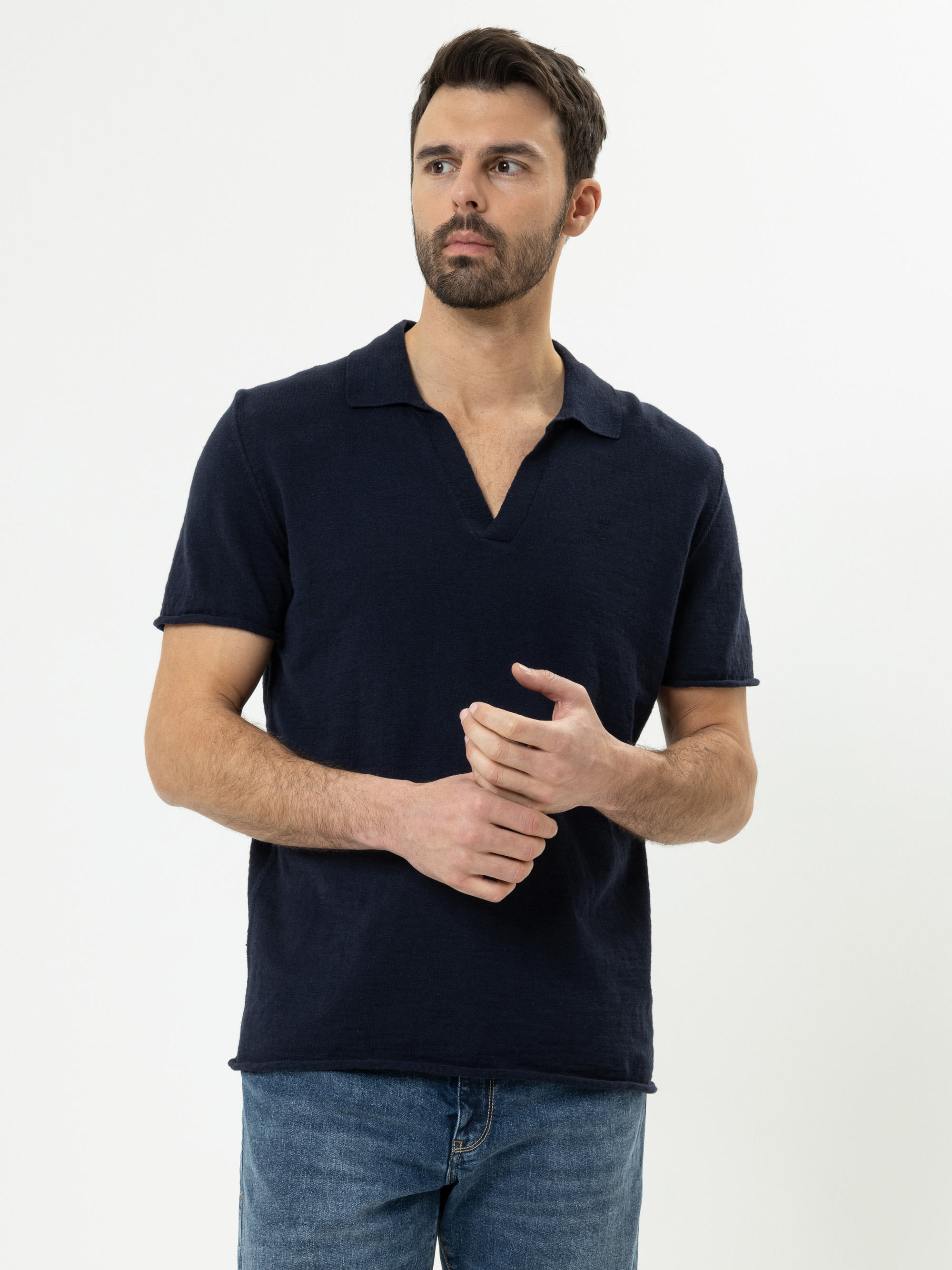 Ein Mann mit dunklem Haar und Bart trägt ein marineblaues, kurzärmeliges Hemd und blaue Jeans. Er steht vor einem schlichten weißen Hintergrund und blickt mit locker gefalteten Händen leicht zur Seite.