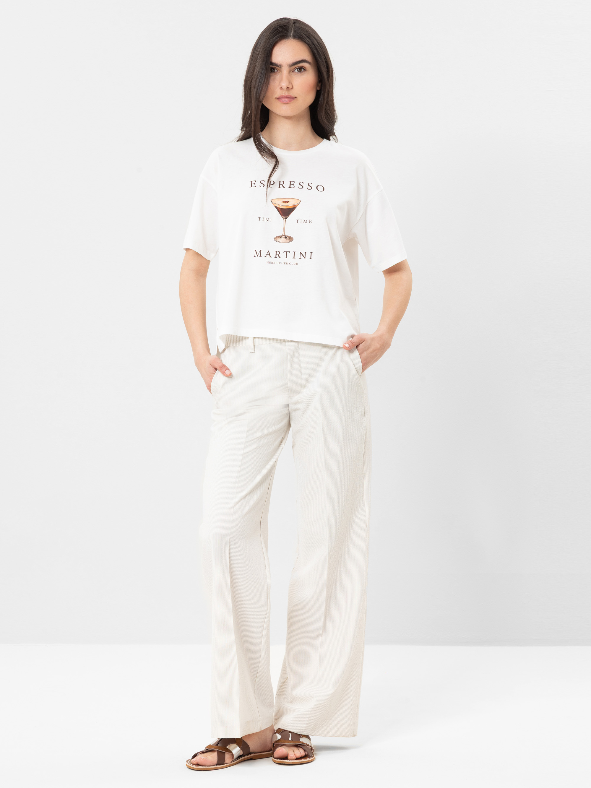 Eine Frau steht vor einem weißen Hintergrund und trägt ein weißes T-Shirt mit Espresso Martini-Text und -Grafik, eine cremefarbene Hose mit weitem Bein und braune Sandalen. Sie hat langes braunes Haar und einen entspannten Gesichtsausdruck.