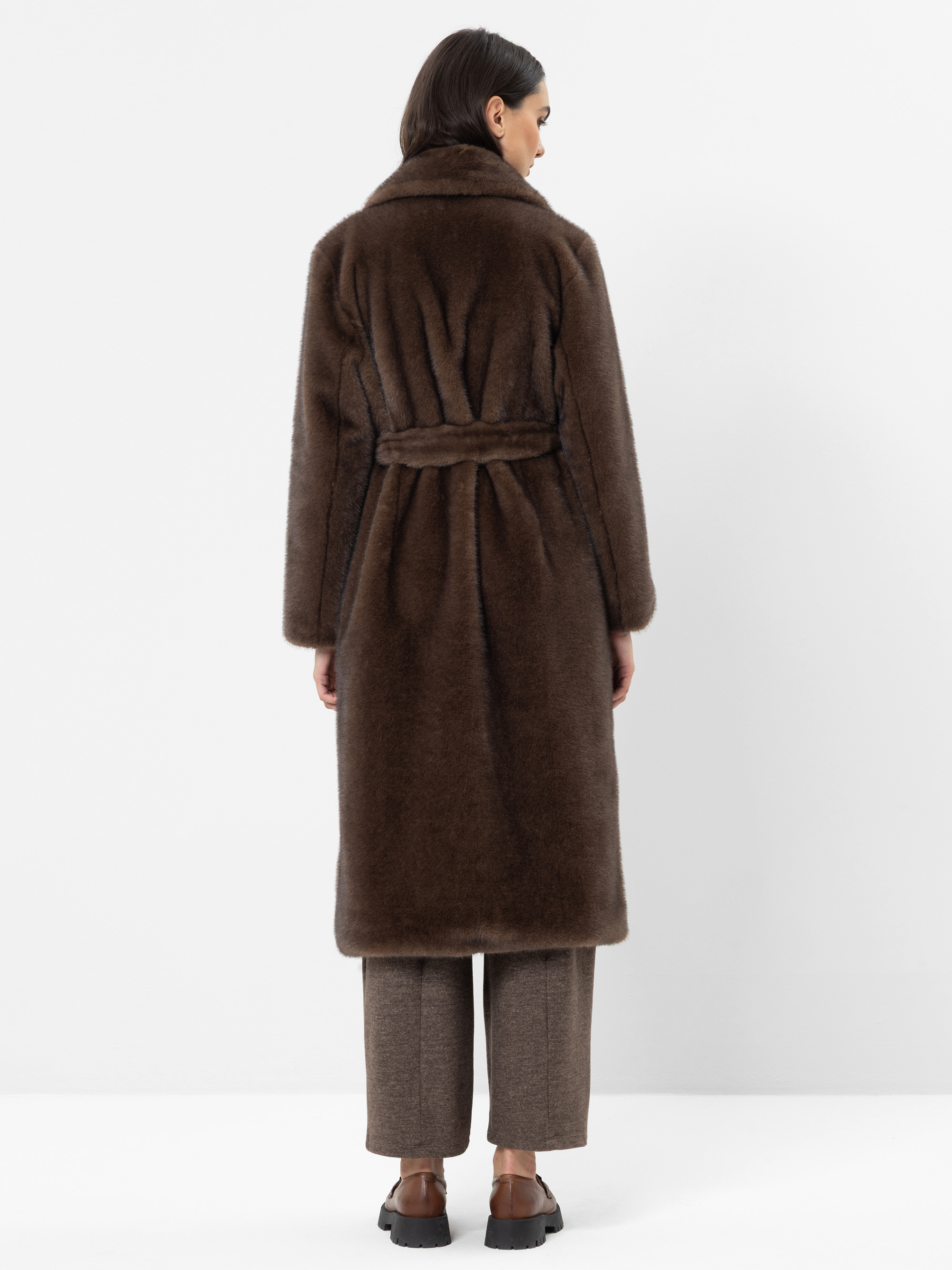 Vor einem schlichten weißen Hintergrund steht eine Person mit dem Rücken zur Kamera und trägt einen langen, dunkelbraunen Pelzmantel mit einem Gürtel in der Taille, eine graue Hose und braune Schuhe.