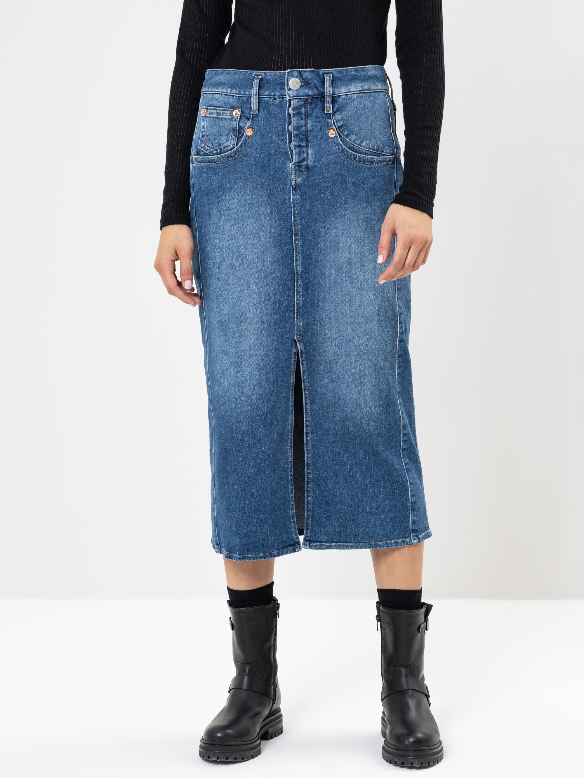 Eine Person, die einen langen blauen Jeansrock mit einem Schlitz an der Vorderseite, ein schwarzes geripptes langärmeliges Oberteil und schwarze Ankle Boots trägt, steht vor einem schlichten weißen Hintergrund.