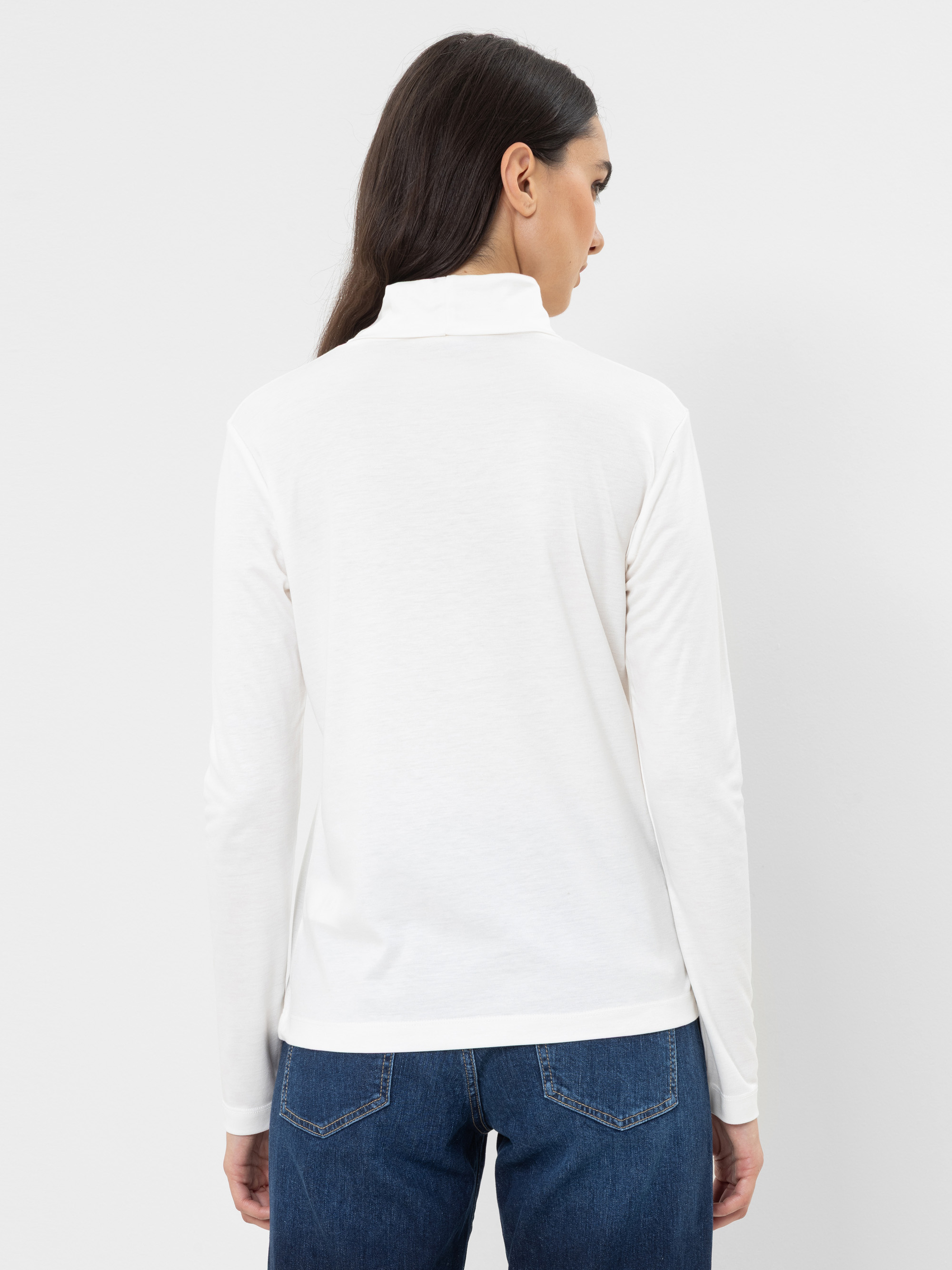 Eine Frau mit langen dunklen Haaren ist von hinten zu sehen; sie trägt ein weißes, langärmeliges Oberteil mit hohem Kragen und eine blaue Jeans und steht vor einem schlichten weißen Hintergrund.