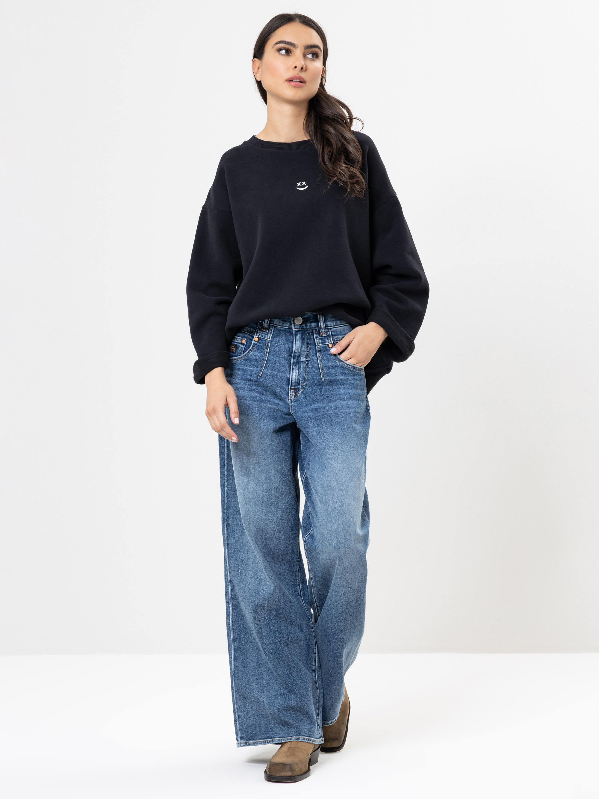 Eine Frau steht vor einem weißen Hintergrund, trägt ein lockeres schwarzes Sweatshirt, weite Bluejeans und braune Schuhe, die linke Hand in der Tasche und den Kopf leicht geneigt.