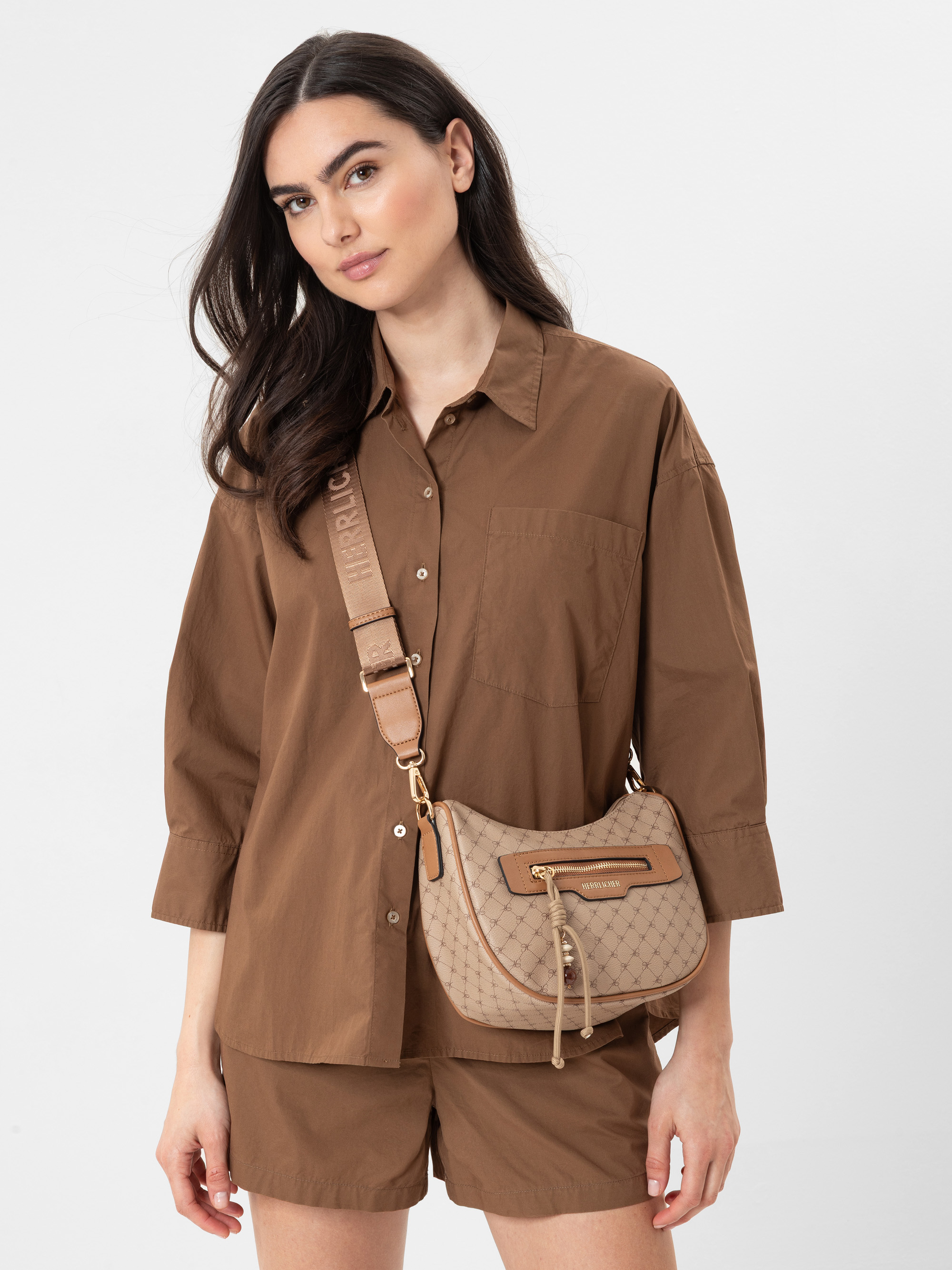 Eine Frau mit langen dunklen Haaren trägt ein lockeres braunes Button-up-Hemd und passende Shorts. Sie trägt eine beigefarbene Umhängetasche mit einem gemusterten Muster und goldenen Akzenten. Sie steht vor einem schlichten, hellen Hintergrund.