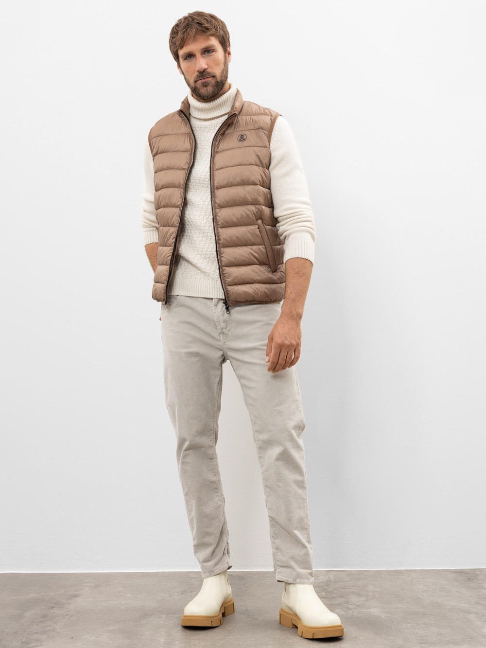 Ein Mann steht vor einer weißen Wand und trägt eine beigefarbene Pufferweste über einem cremefarbenen Rollkragenpullover, eine hellgraue Hose und cremefarbene Stiefel. Er hat eine entspannte Haltung, eine Hand an der Seite und die andere in der Tasche.