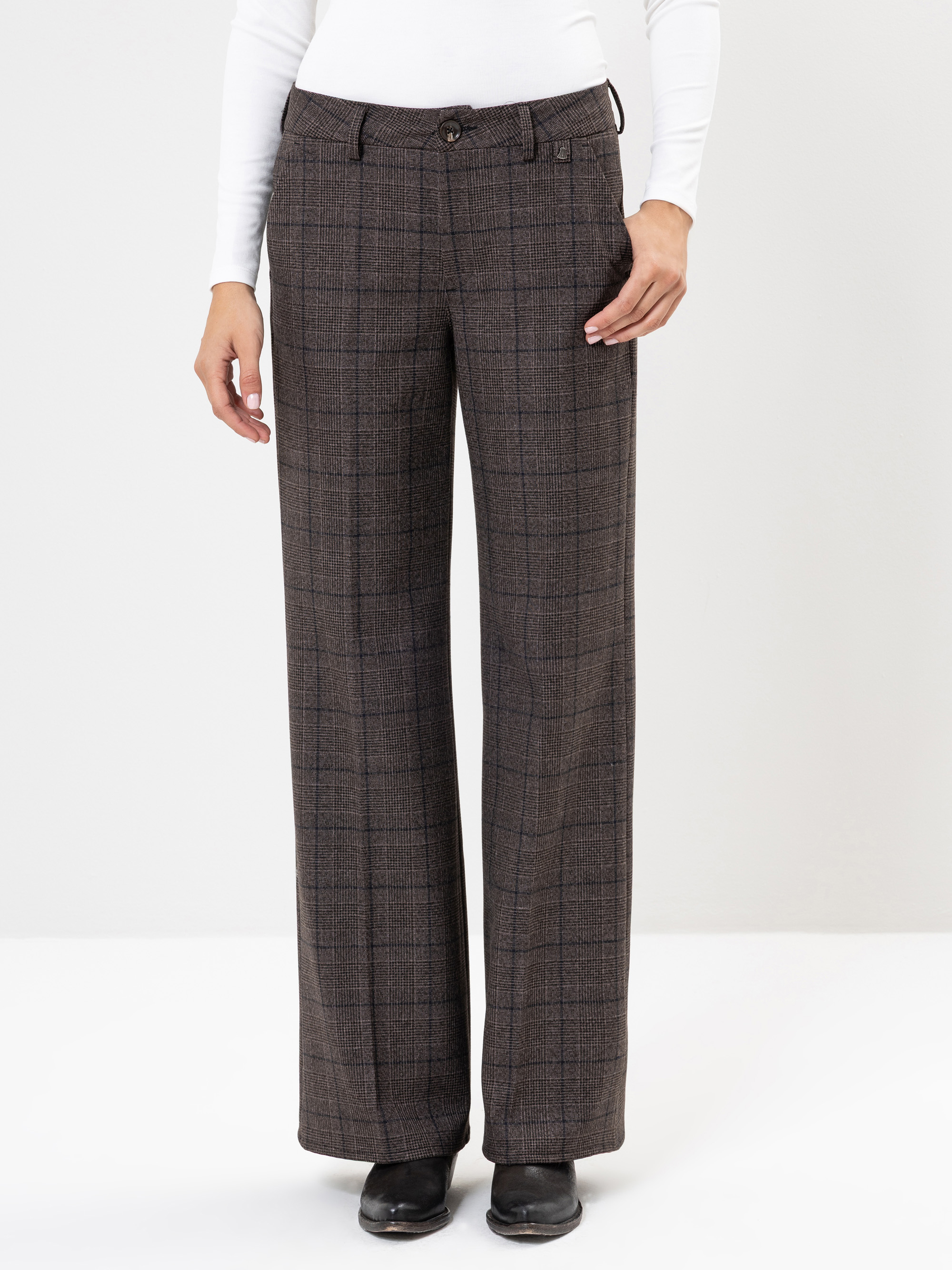 Eine Person, die eine braun karierte Hose mit weitem Bein, ein weißes langärmeliges Oberteil und schwarze Schuhe trägt, steht vor einem schlichten weißen Hintergrund.