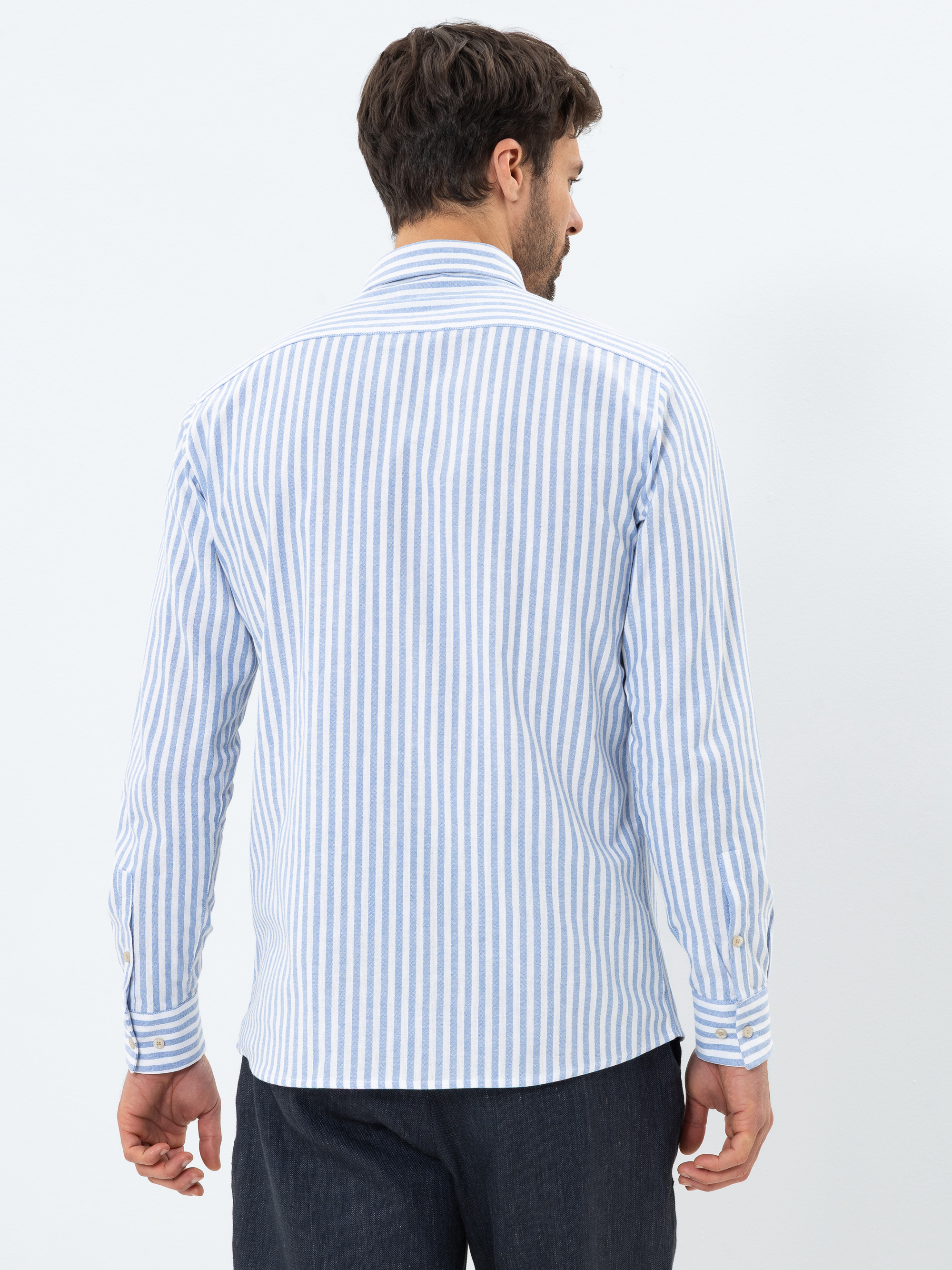 Ein Mann mit kurzen braunen Haaren ist von hinten zu sehen. Er trägt ein blau-weiß vertikal gestreiftes langärmeliges Hemd und eine dunkle Hose und steht vor einem schlichten hellen Hintergrund.