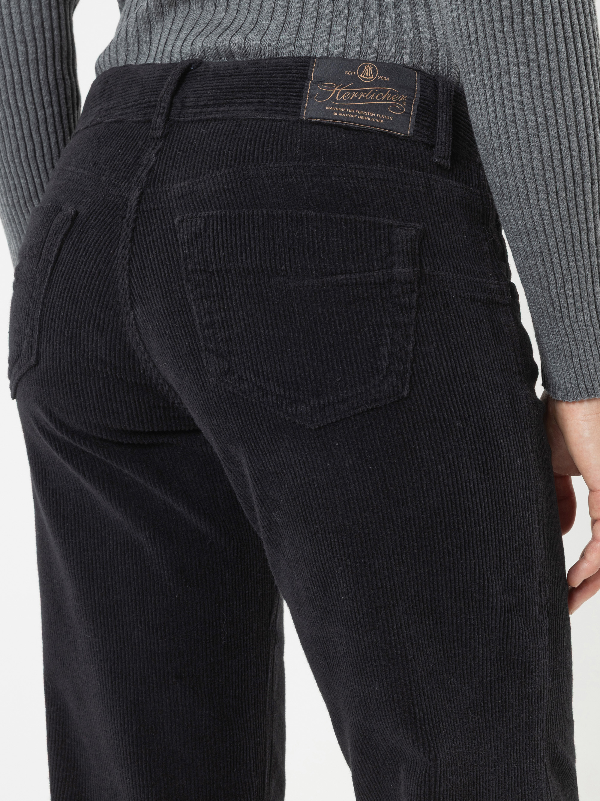Eine Person, die eine schwarze Cordhose und einen grauen gerippten Pullover trägt, von hinten ab der Taille abgebildet. Die Hose hat zwei Gesäßtaschen und ein braunes Etikett am Bund.