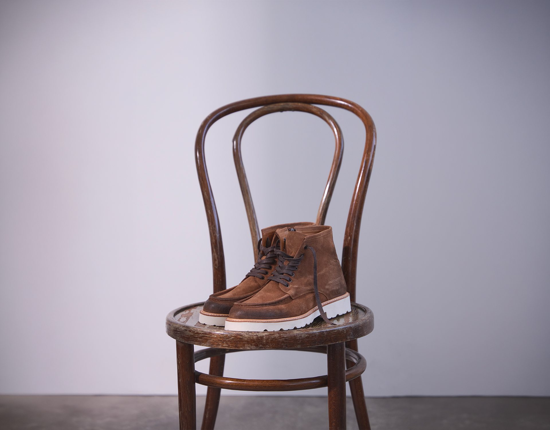 Ein Paar braune Wildlederstiefel mit weißen Sohlen steht auf der Sitzfläche eines alten Holzstuhls vor einem schlichten, hellgrauen Hintergrund.