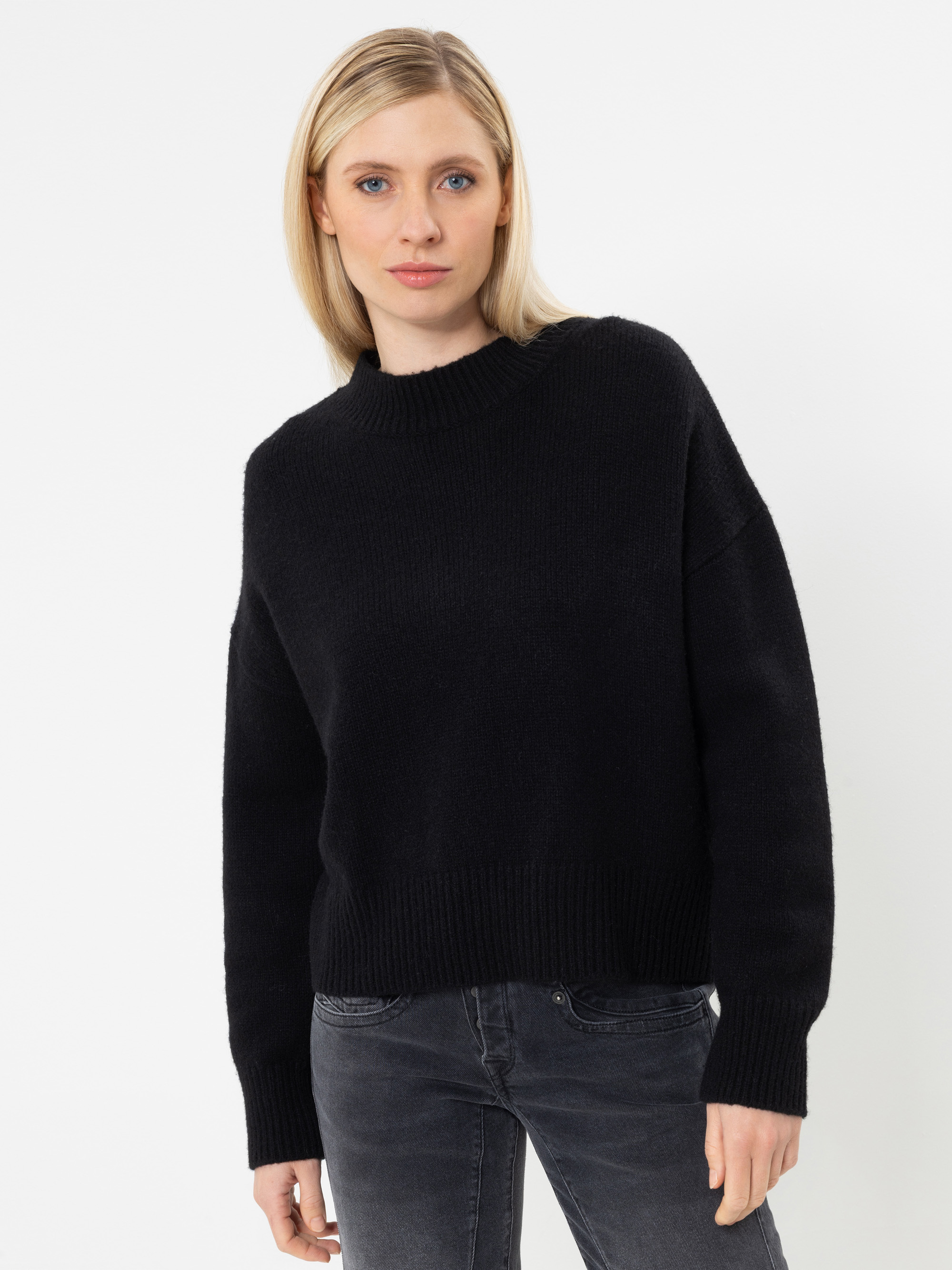 Eine Frau mit glattem blondem Haar trägt einen schwarzen, langärmeligen Pullover und eine dunkle Jeans und steht vor einem schlichten weißen Hintergrund.