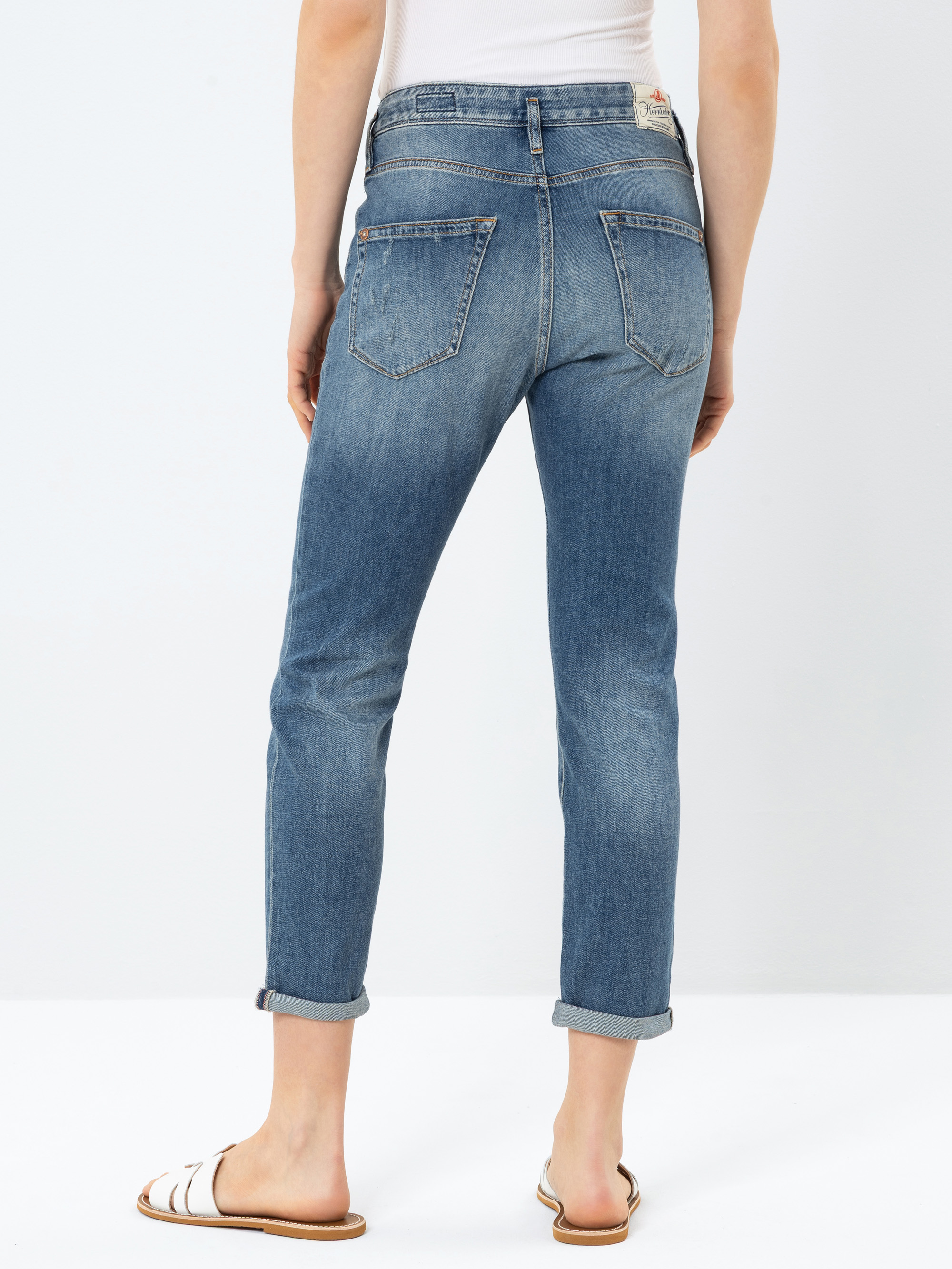 Eine Person, die blaue Jeans mit Bündchen und weiße Sandalen trägt, wird von hinten gezeigt und steht vor einem schlichten weißen Hintergrund. Die Person trägt außerdem ein weißes Oberteil.