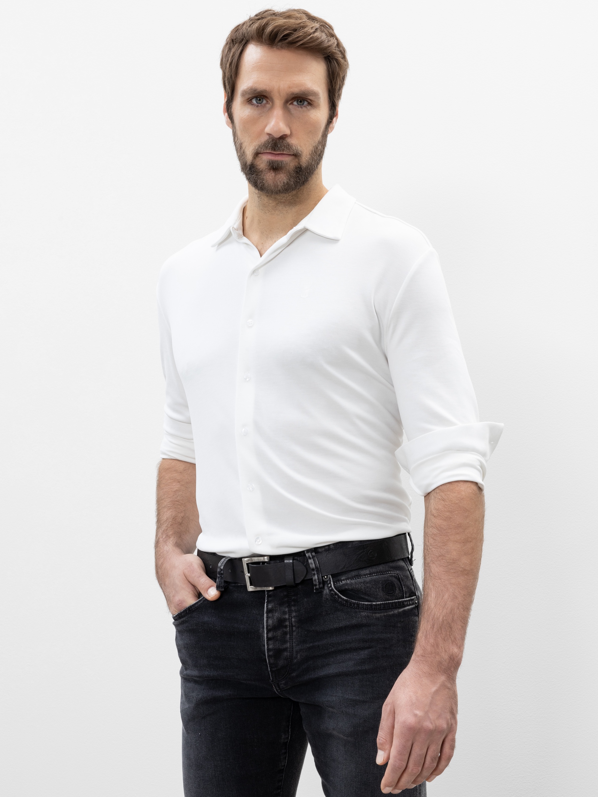 Ein Mann mit hellbraunem Haar und Bart steht vor einem weißen Hintergrund und trägt ein weißes Button-up-Hemd mit hochgekrempelten Ärmeln, schwarze Jeans und einen schwarzen Gürtel. Er hat eine Hand in der Tasche und einen neutralen Gesichtsausdruck.
