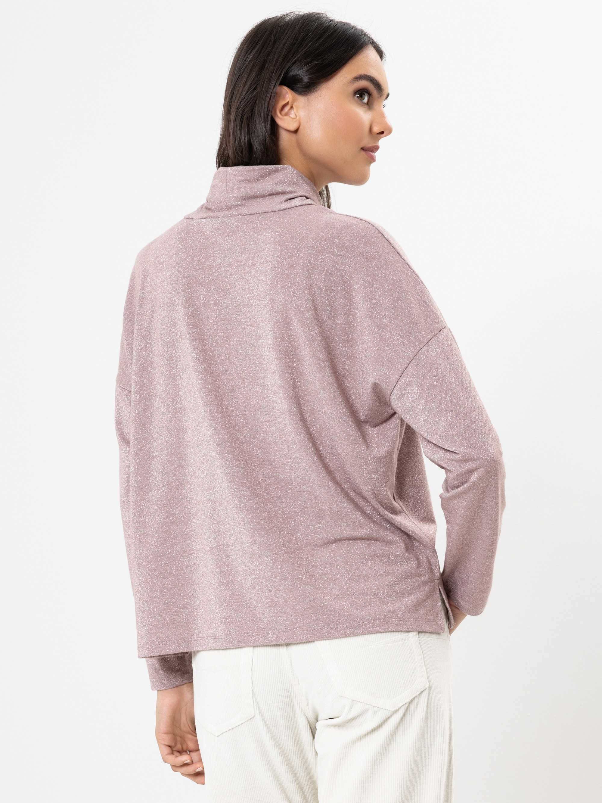 Frau mit dunklem Haar, die einen hellvioletten Rollkragenpullover und eine weiße Hose trägt, steht vor einem weißen Hintergrund und blickt über ihre Schulter, wobei sie die Rückseite ihres Outfits zeigt.