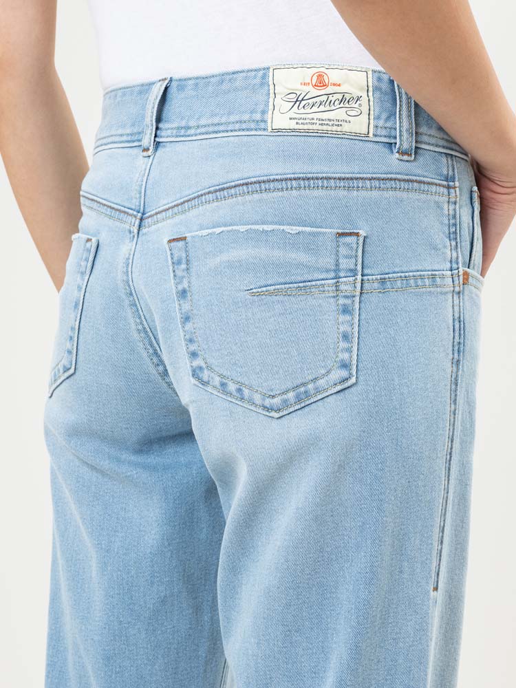 Person trägt eine hellblaue Jeans mit einem weißen "Herrlicher"-Etikett am Bund. Das Bild konzentriert sich auf die Rückseite der Jeans und zeigt die Taschen und einen Teil der Hand der Person in der Tasche.