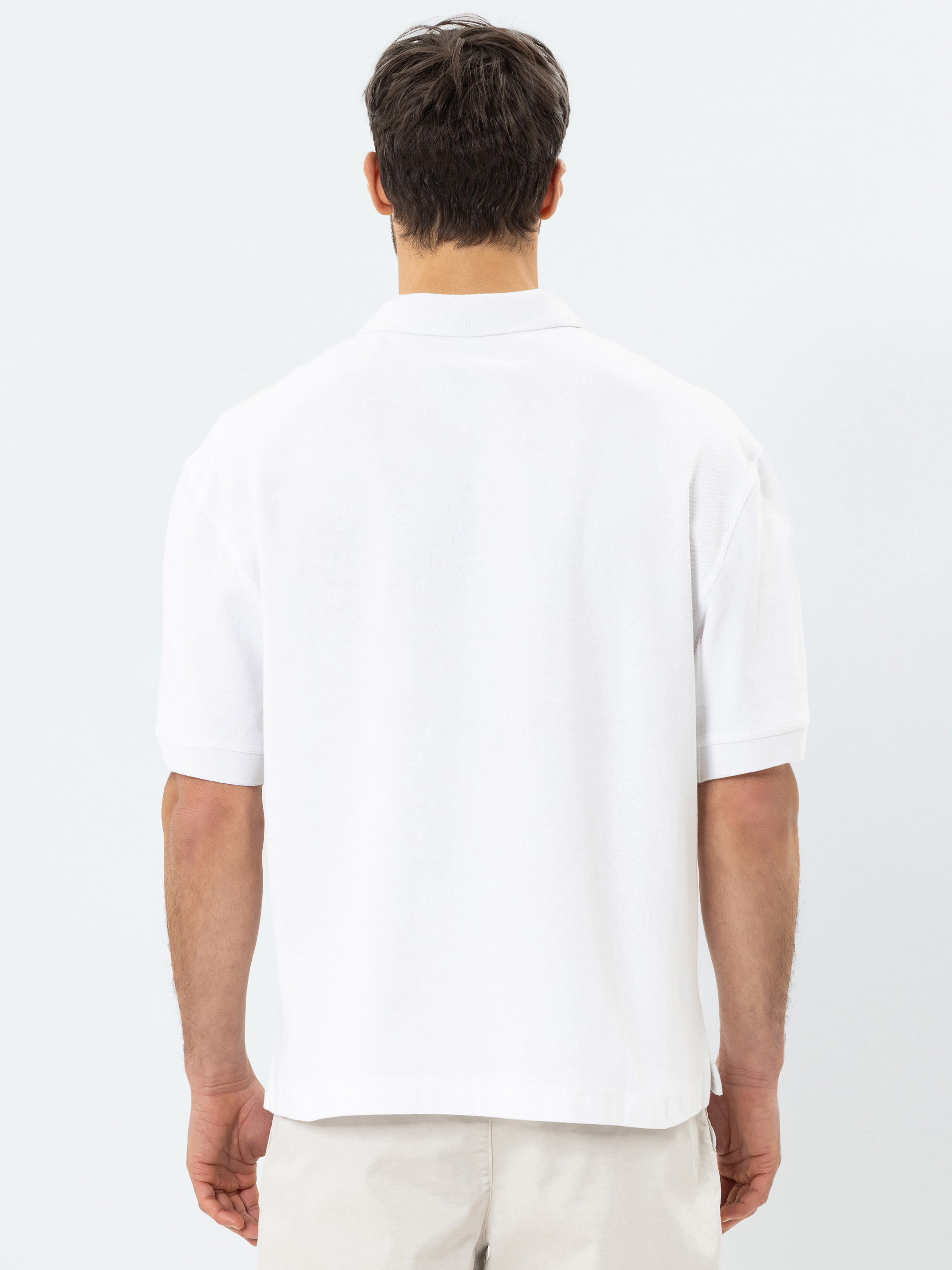 Ein Mann steht mit dem Rücken zur Kamera und trägt ein schlichtes weißes, kurzärmeliges Polohemd und helle Shorts vor einem schlichten, hellen Hintergrund.