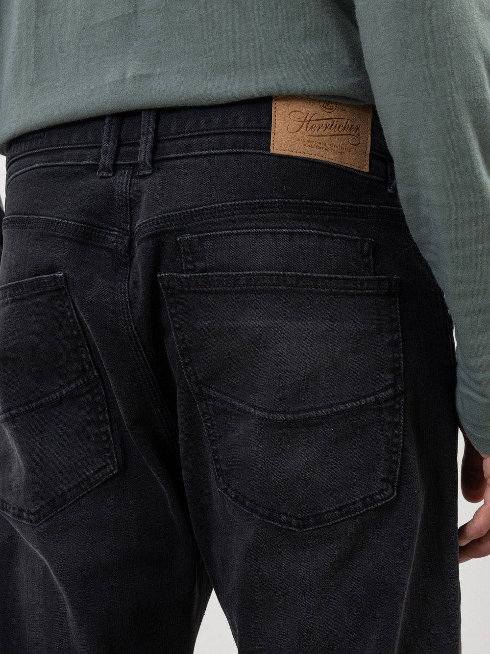 Eine Person, die eine schwarze Jeans mit großen Gesäßtaschen und einem hellbraunen Lederaufnäher am Bund trägt, dazu ein grünes langärmeliges Hemd.