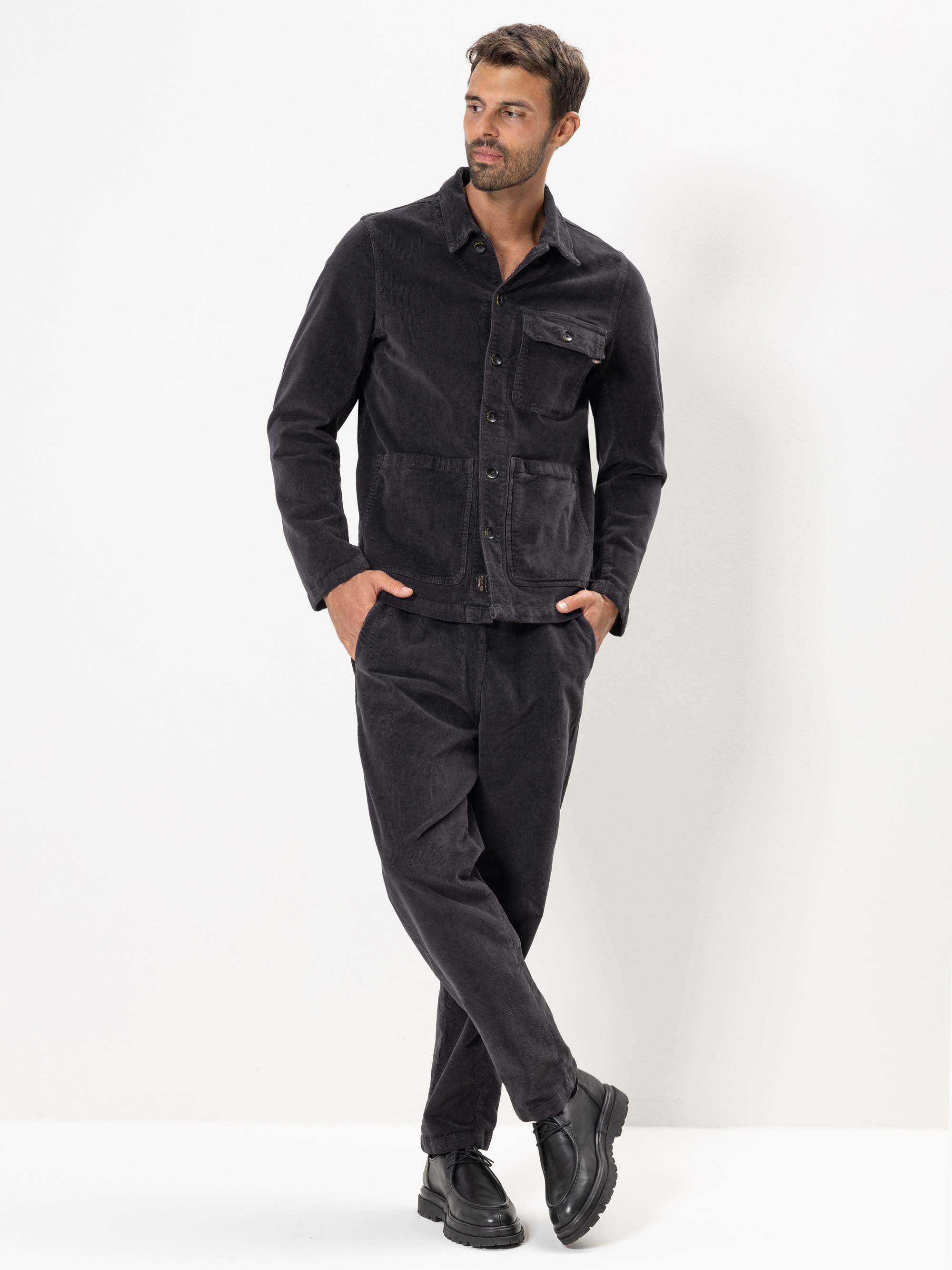 Ein Mann steht vor einem weißen Hintergrund und trägt eine dunkle Cordjacke und eine passende Hose mit schwarzen Schuhen. Er blickt zur Seite, hat ein Bein vor dem anderen gekreuzt und eine Hand in der Tasche.