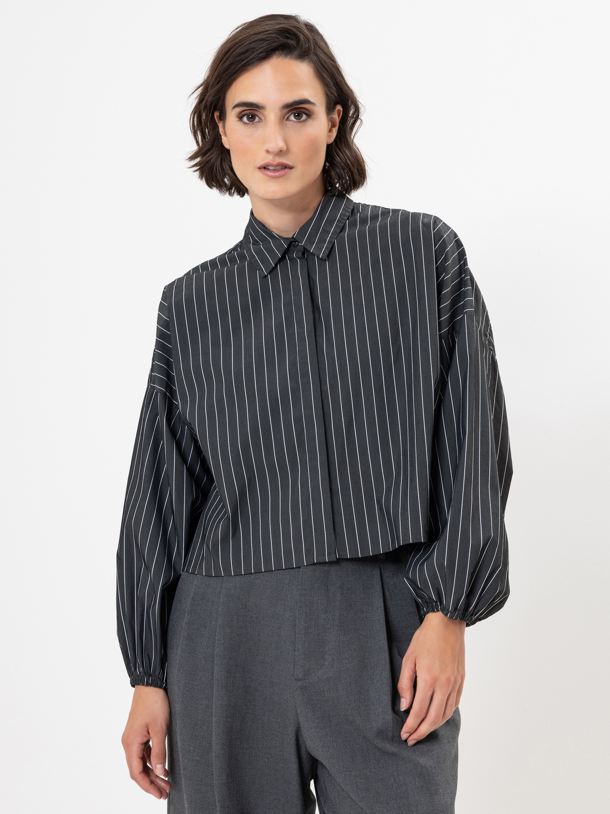 Eine Person mit kurzen braunen Haaren trägt eine locker sitzende, langärmelige, schwarze Bluse mit weißen Längsstreifen und eine graue Hose mit hoher Taille vor einem schlichten weißen Hintergrund.