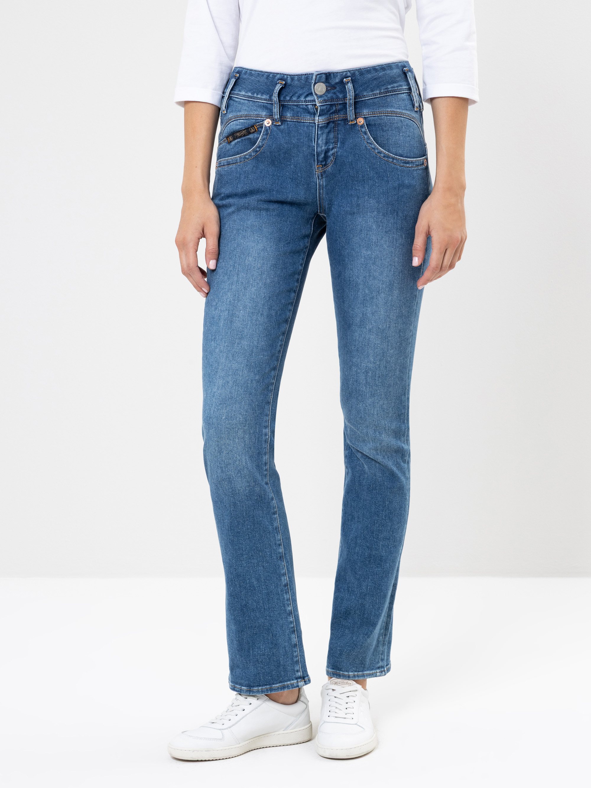 Person, die mittelblaue Jeans mit geradem Bein, ein weißes langärmeliges Oberteil und weiße Turnschuhe trägt und vor einem schlichten hellen Hintergrund steht. Das Bild ist von den Schultern bis zu den Füßen abgeschnitten.