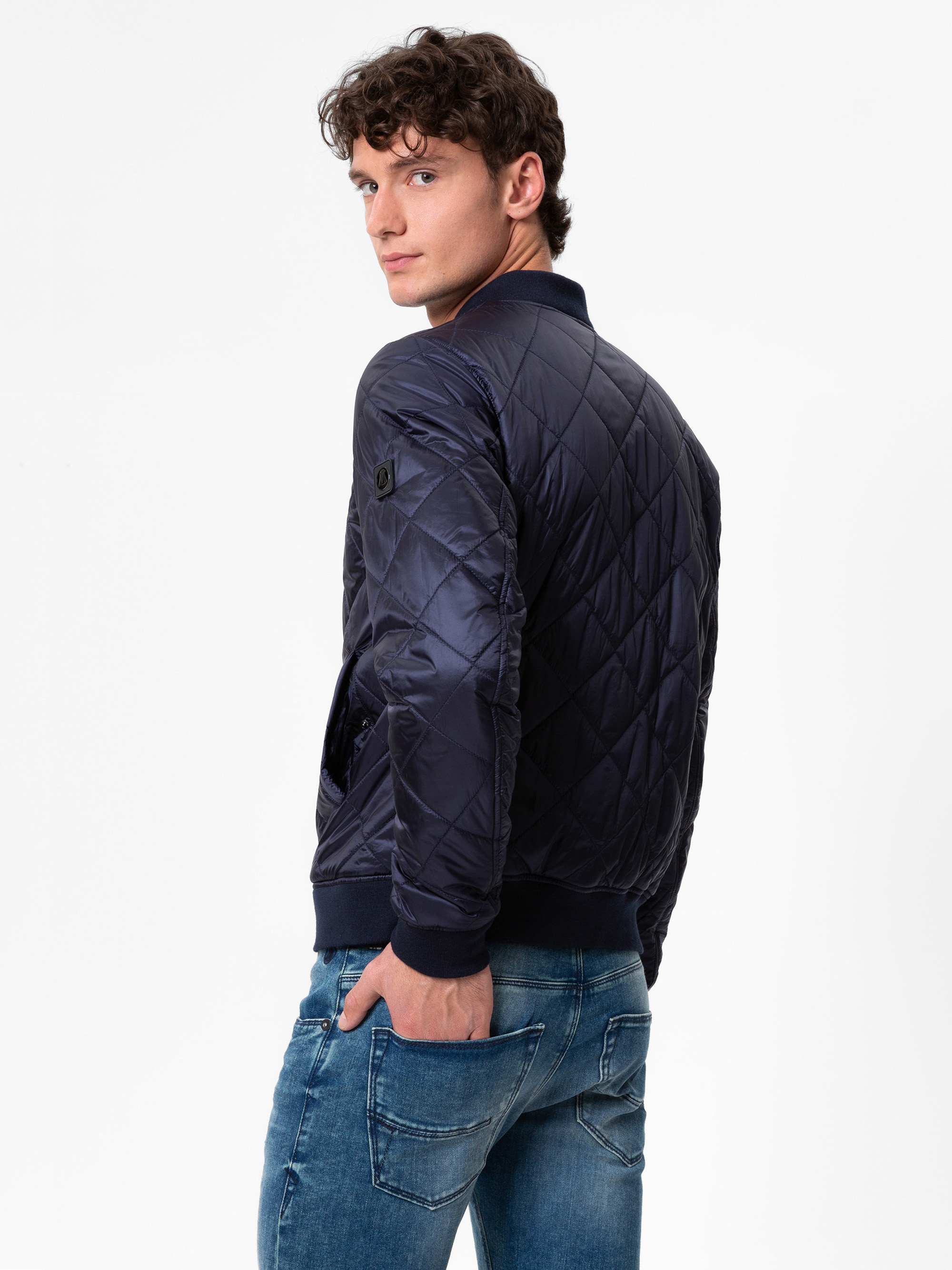 Ein junger Mann mit lockigen braunen Haaren, der eine dunkle Steppjacke und blaue Jeans trägt, steht mit dem Gesicht zur Seite und blickt über seine Schulter auf einen schlichten weißen Hintergrund.