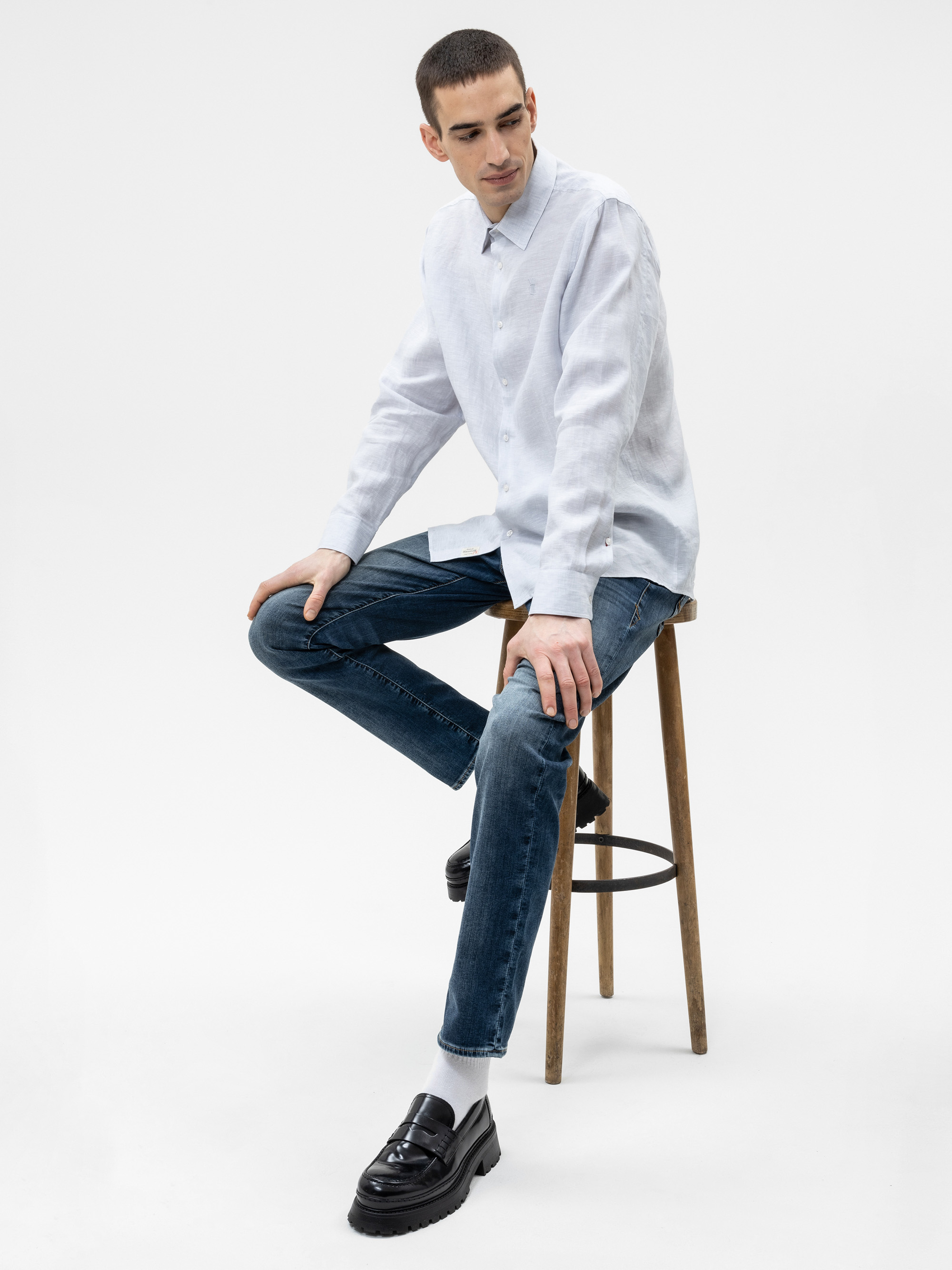 Ein Mann mit kurzen dunklen Haaren sitzt auf einem Holzhocker vor einem schlichten weißen Hintergrund. Er trägt ein helles Button-up-Hemd, blaue Jeans, weiße Socken und schwarze Halbschuhe.