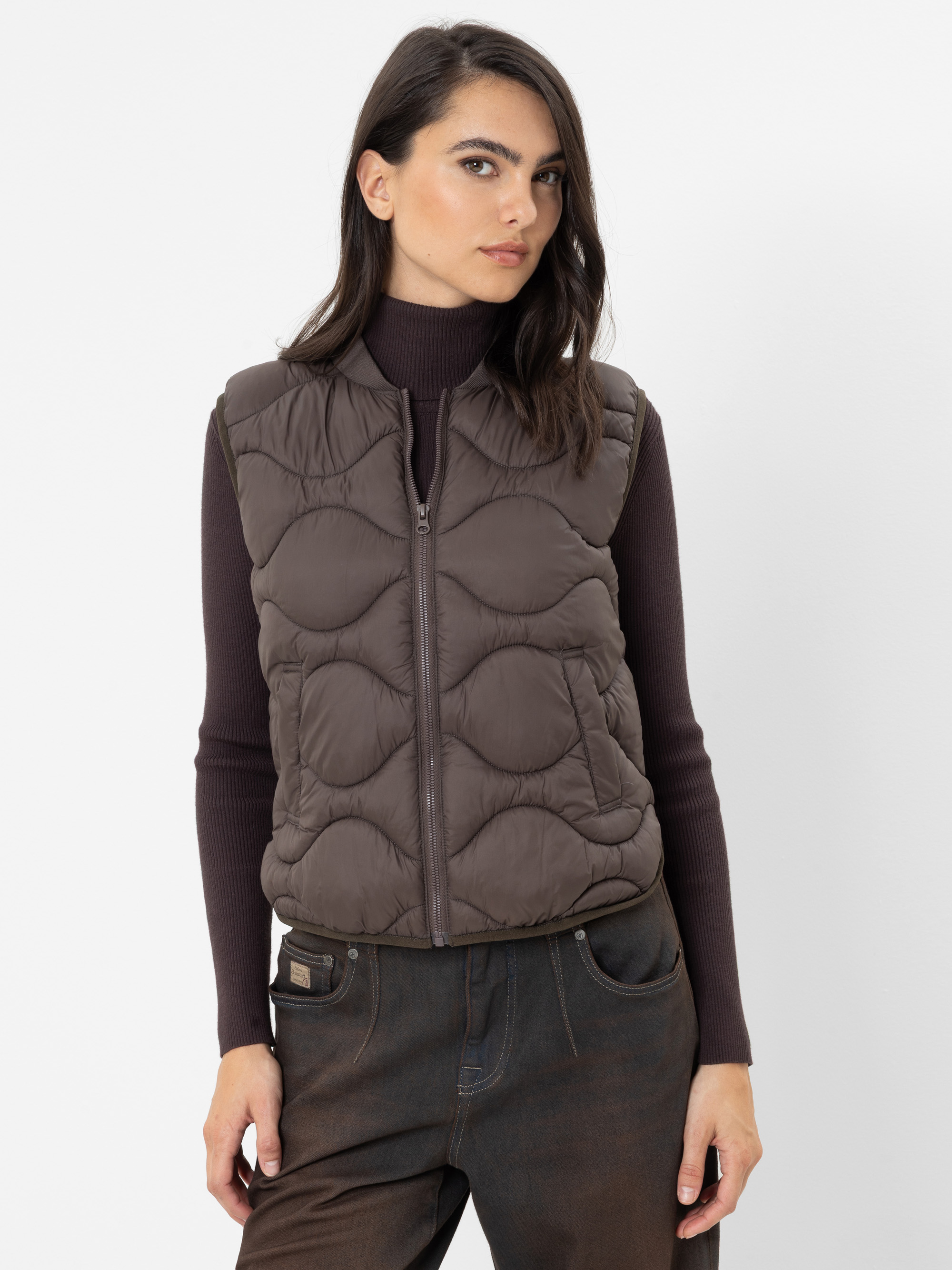 Eine Frau, die eine braune, gesteppte Weste über einem dunklen Rollkragenpullover und dunklen Jeans trägt, steht vor einem schlichten weißen Hintergrund und blickt mit ausdruckslosem Gesicht leicht zur Seite.