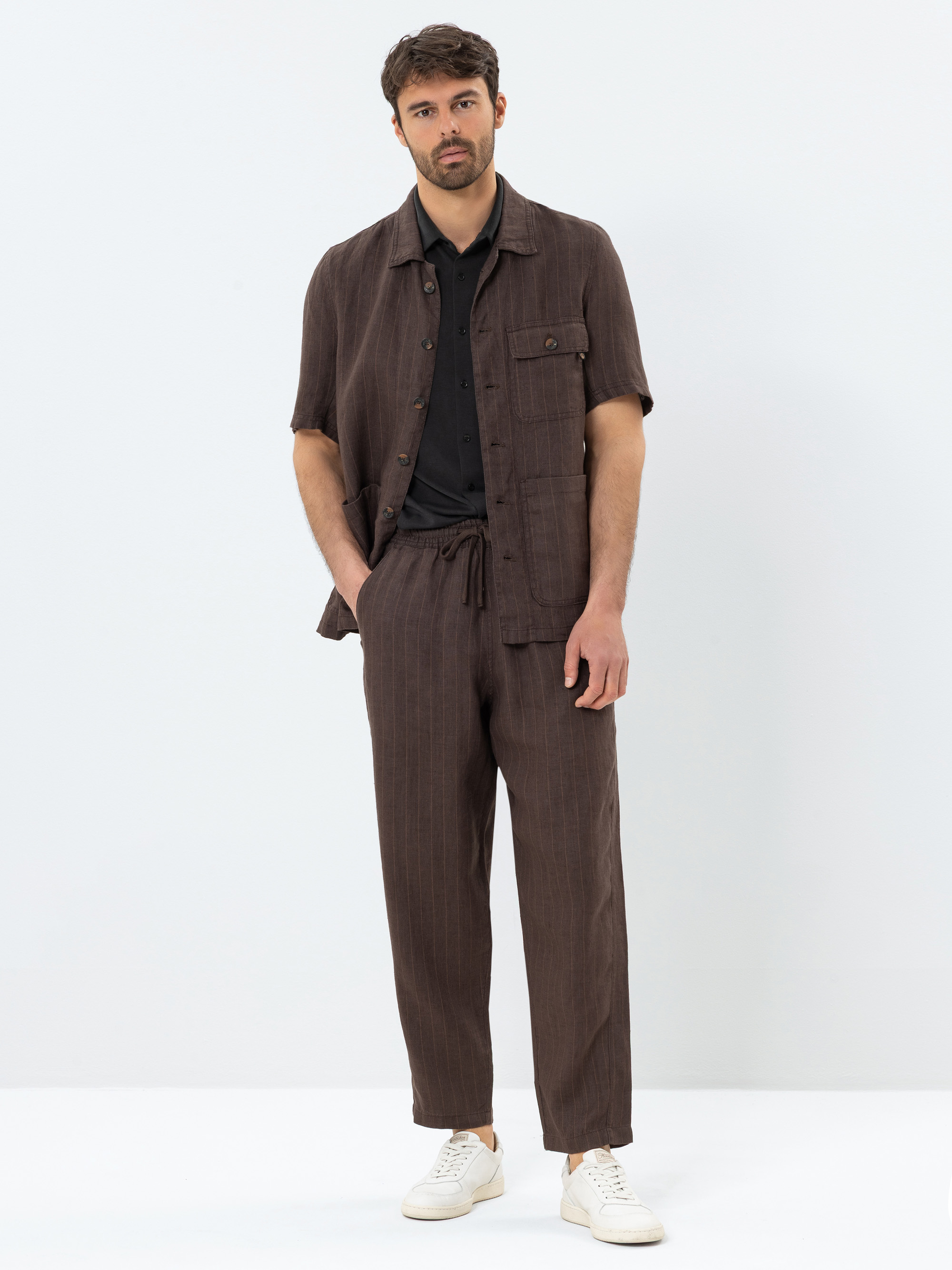 Ein Mann steht vor einem schlichten Hintergrund und trägt ein braunes, kurzärmeliges Button-up-Hemd mit einer Brusttasche, eine dazu passende braune Hose mit Kordelzug, darunter ein schwarzes Hemd und weiße Turnschuhe.