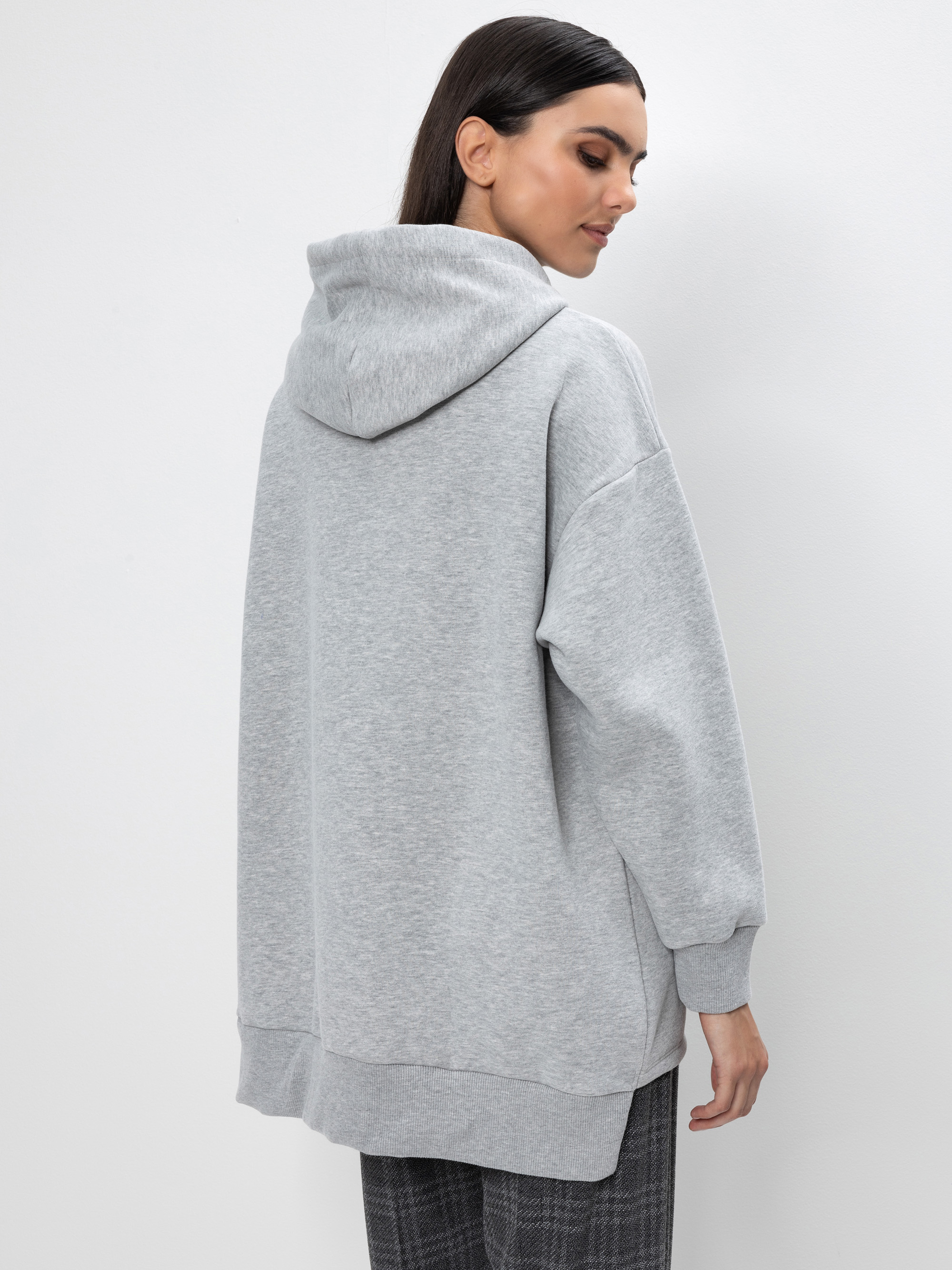 Eine Frau mit langen, dunklen Haaren steht mit dem Gesicht zur Wand, trägt einen übergroßen hellgrauen Kapuzenpulli und eine grau karierte Hose und steht vor einem schlichten weißen Hintergrund.