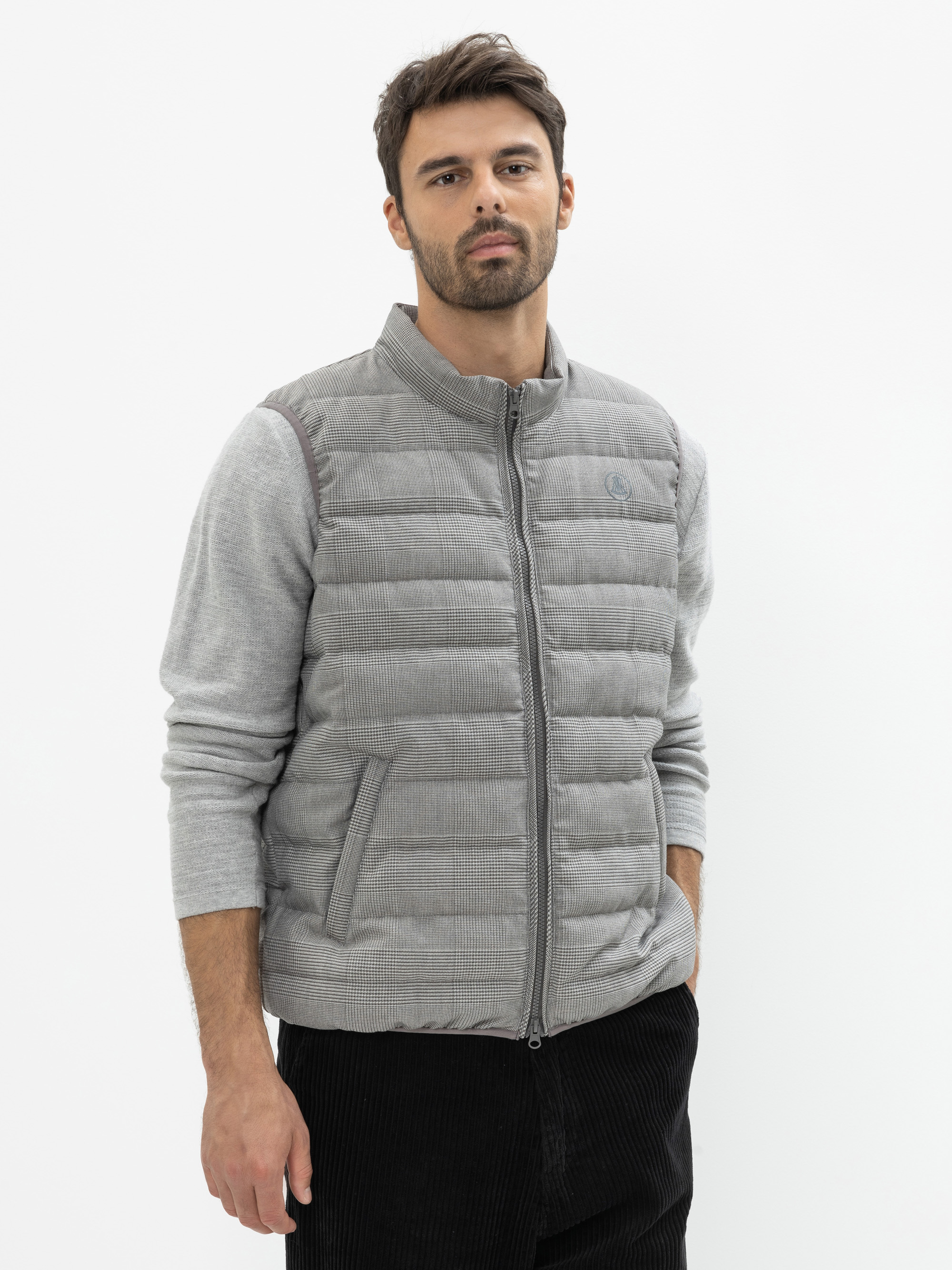 Ein Mann mit kurzen dunklen Haaren und einem Bart trägt eine hellgraue Steppweste über einem grauen langärmeligen Hemd und eine schwarze Hose und steht vor einem schlichten weißen Hintergrund.