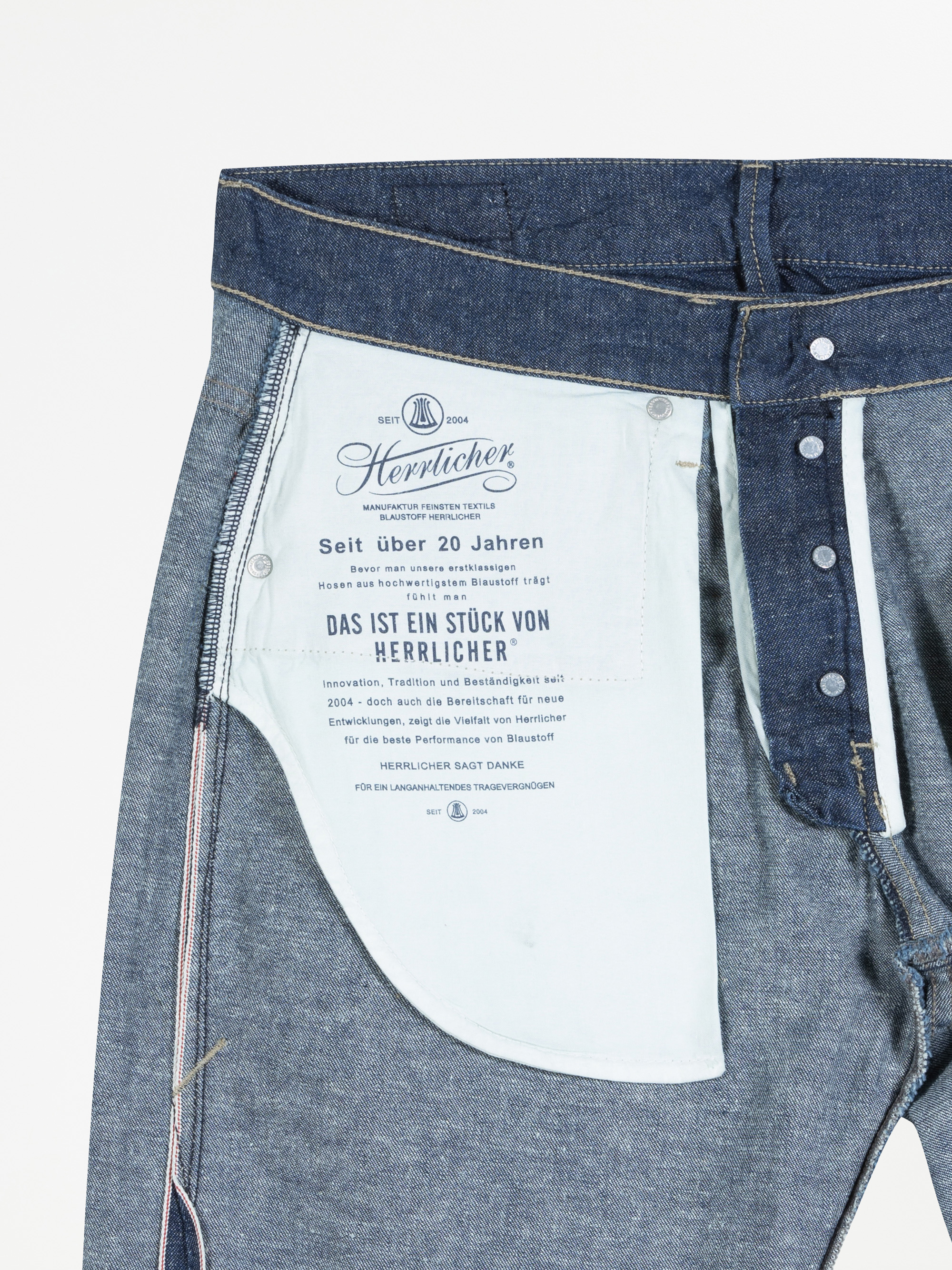 Nahaufnahme der vorderen Innentasche einer Jeans mit blauem Stoff, sichtbaren Nähten, Knöpfen und einem gedruckten Etikett mit deutschem und englischem Text der Marke Herrlicher.