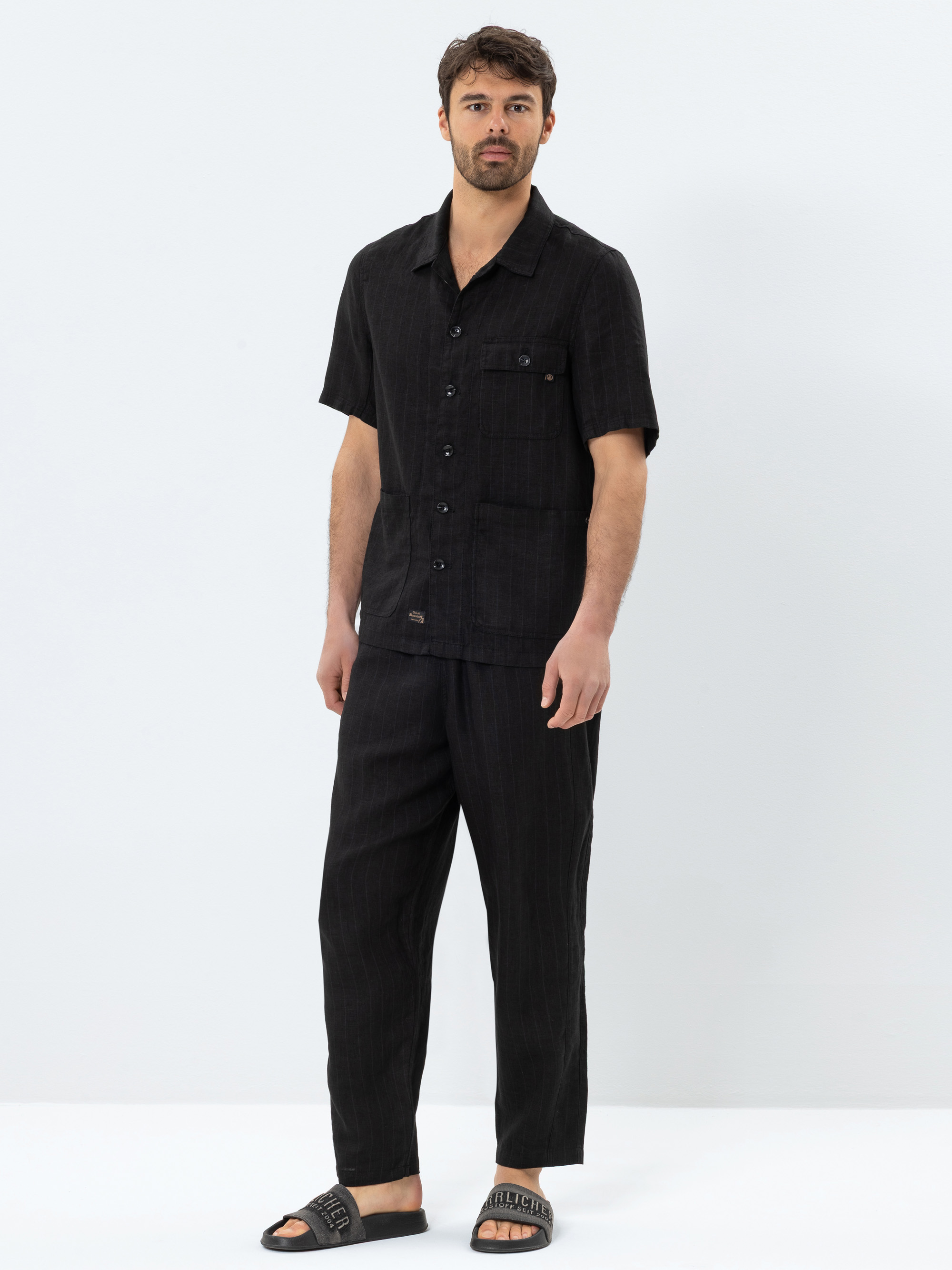 Ein Mann steht vor einem schlichten weißen Hintergrund und trägt ein schwarzes, kurzärmeliges Button-up-Hemd und eine passende schwarze Hose. Er hat kurzes braunes Haar, einen Bart und trägt schwarze Slide-Sandalen.