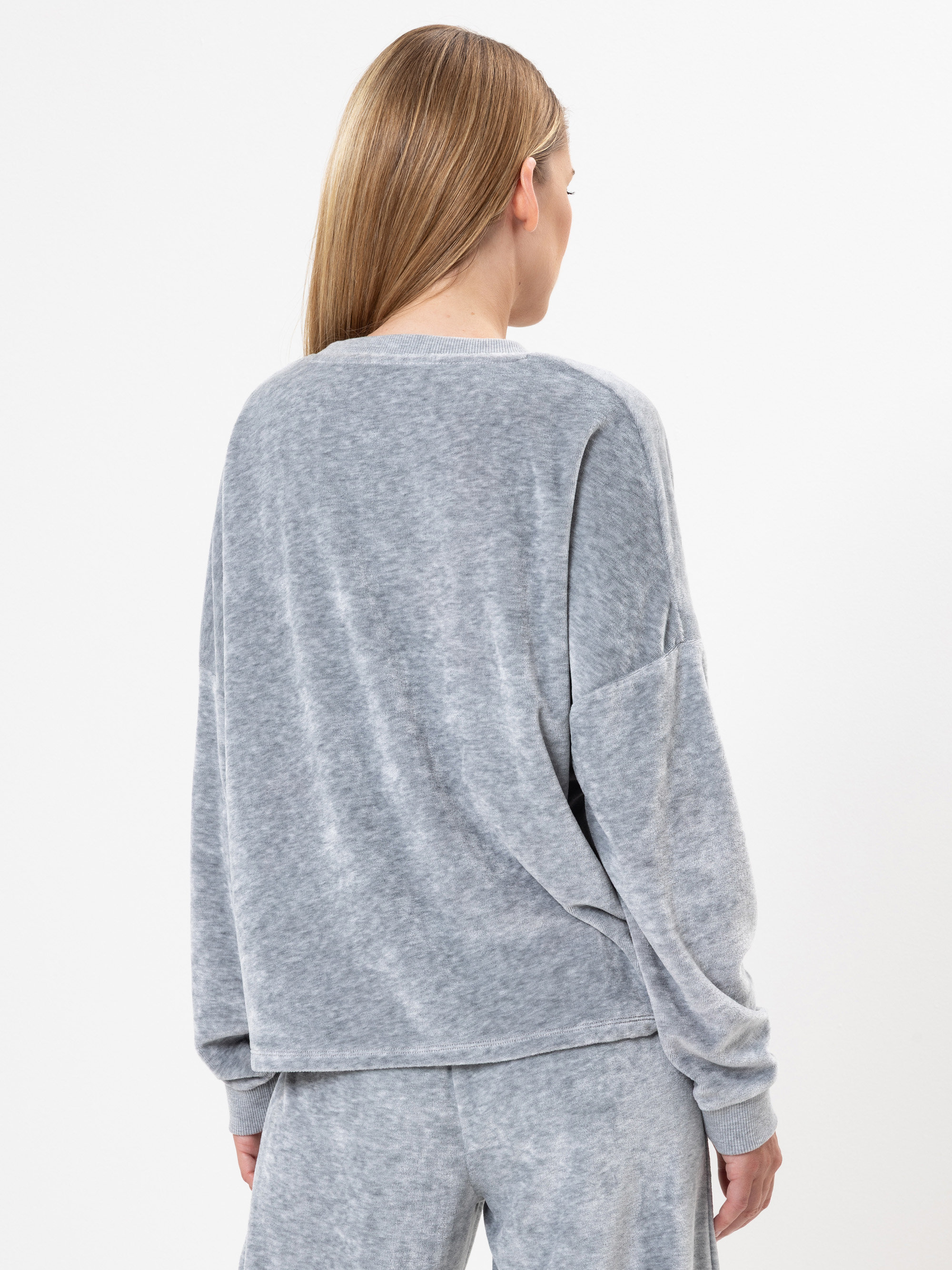 Eine Person mit glattem, hellbraunem Haar wird von hinten gezeigt. Sie trägt einen locker sitzenden, langärmeligen grauen Pullover und eine dazu passende graue Hose vor einem schlichten weißen Hintergrund.