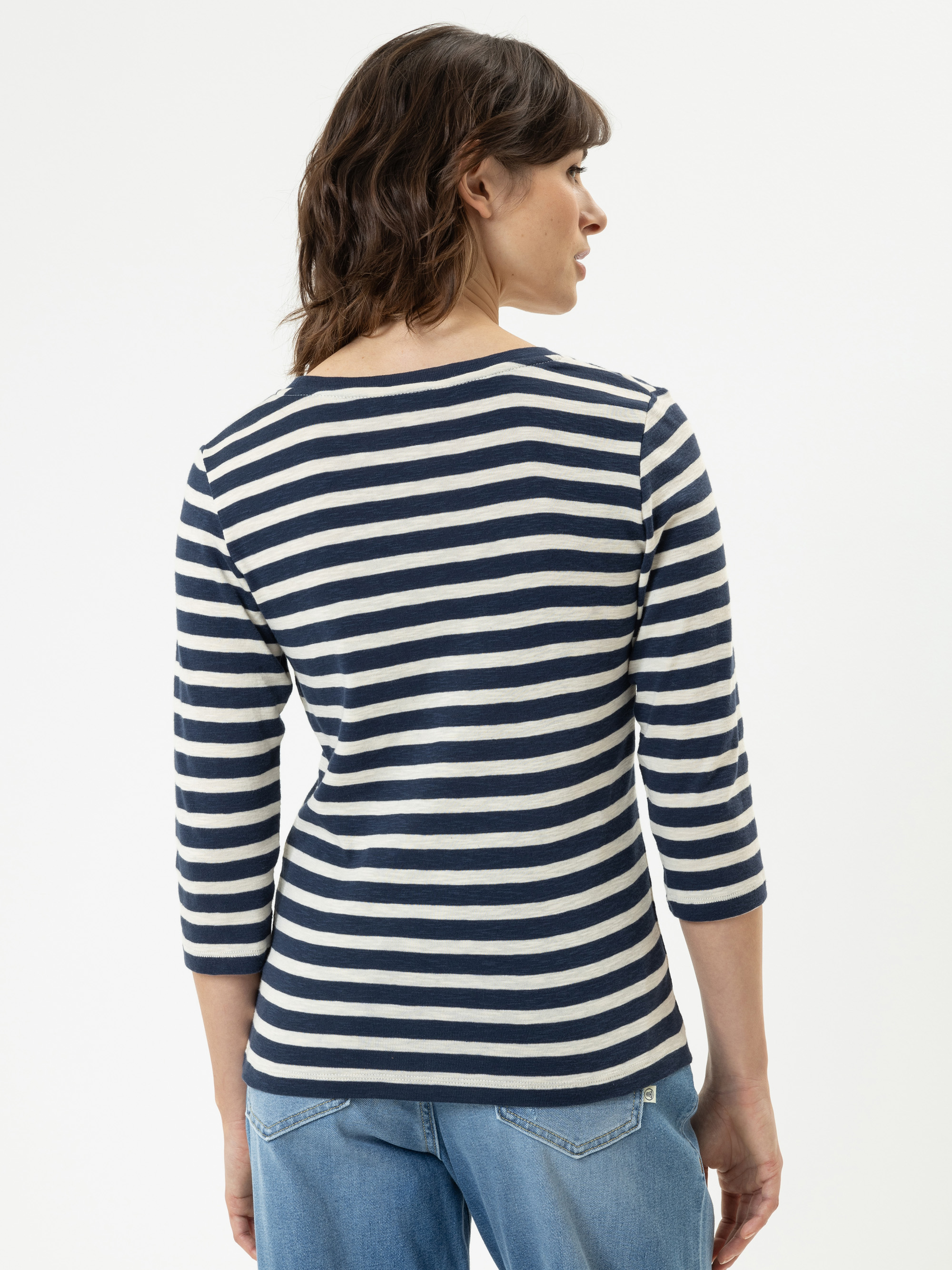 Eine Person mit braunem, gewelltem Haar ist von hinten zu sehen. Sie trägt ein marineblau-weiß gestreiftes, dreiviertelärmeliges Oberteil und hellblaue Jeans vor einem schlichten weißen Hintergrund.