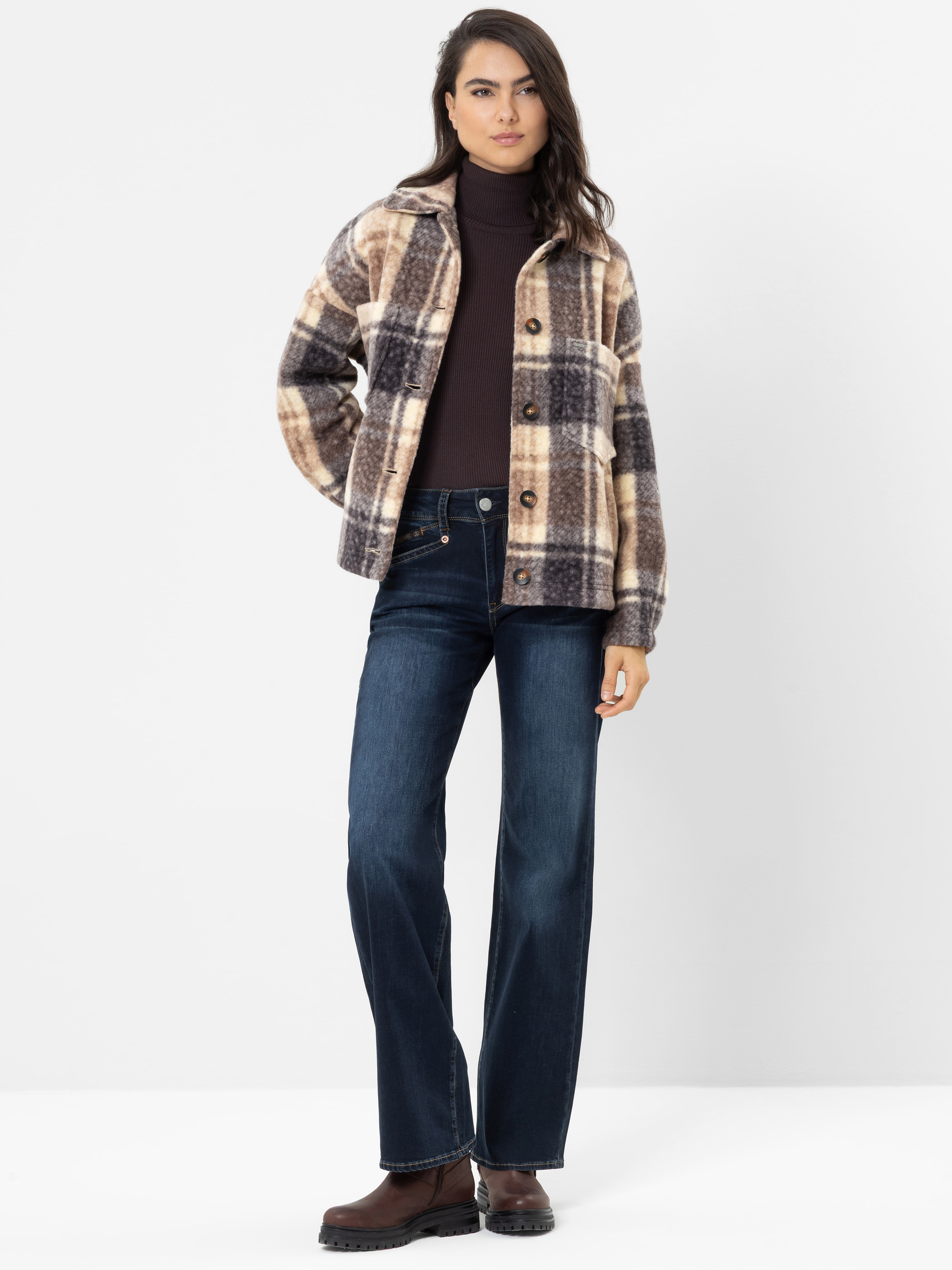 Eine Frau steht vor einem weißen Hintergrund und trägt eine braune karierte Jacke, einen dunklen Rollkragenpullover, dunkelblaue Jeans und braune Stiefel. Eine Hand steckt in ihrer Tasche, und sie hat langes dunkles Haar.