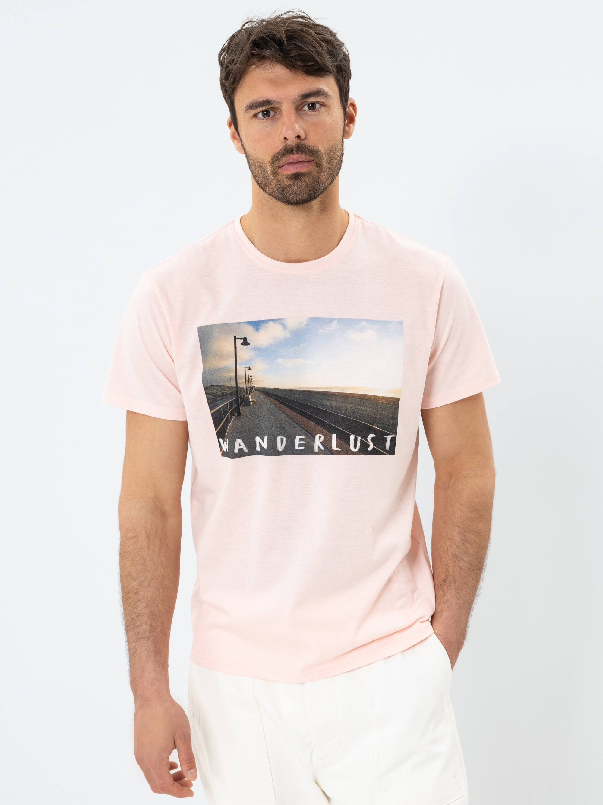Ein Mann mit dunklen Haaren und einem Bart trägt ein hellrosa T-Shirt mit dem Foto einer Strandpromenade und dem Aufdruck WANDERLUST. Er steht vor einem schlichten weißen Hintergrund und schaut ernst.