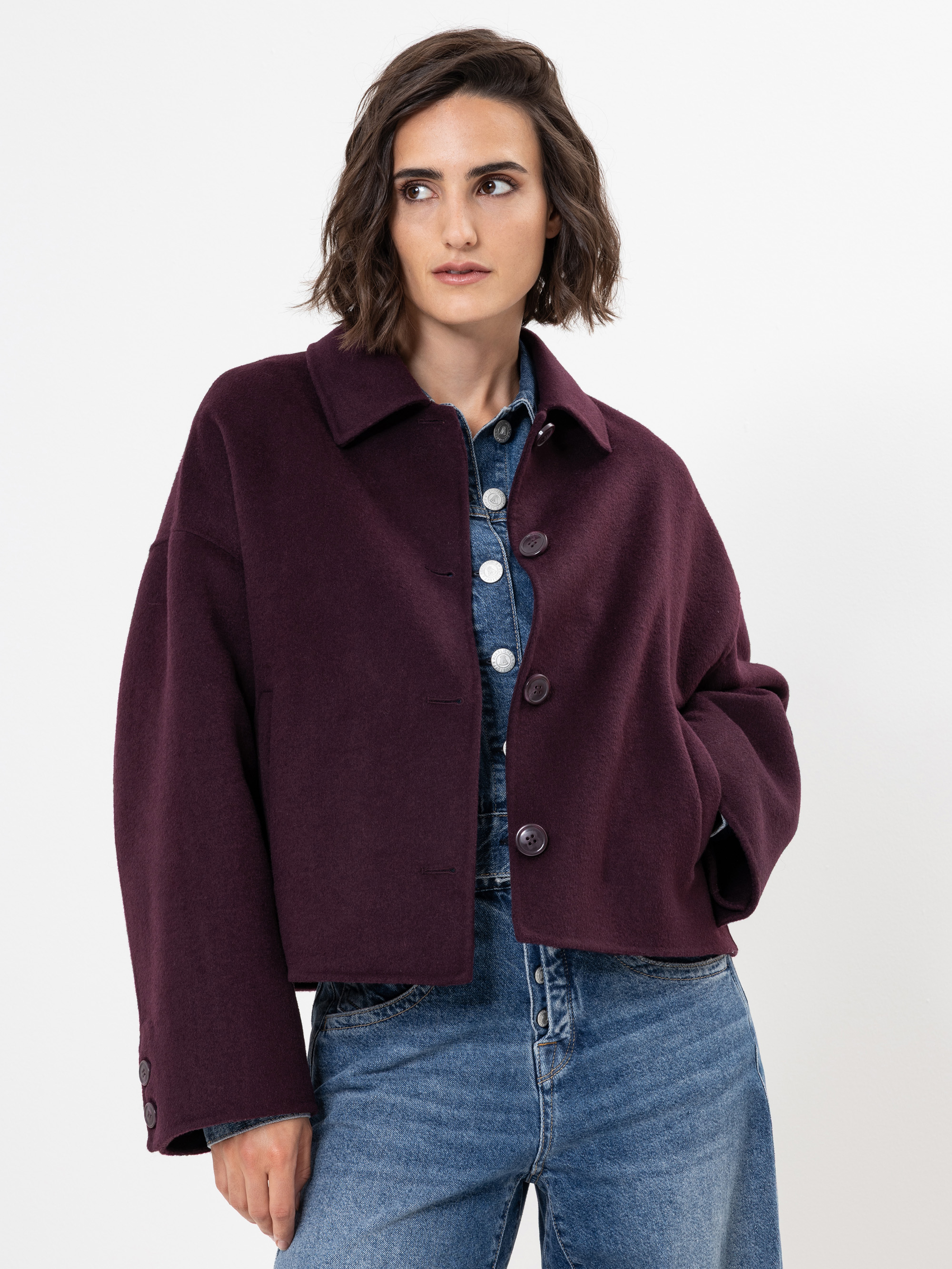 Eine Frau mit kurzen braunen Haaren trägt eine abgeschnittene, dunkelviolette Jacke über einem blauen Jeanshemd und blauen Jeans und steht vor einem schlichten weißen Hintergrund.