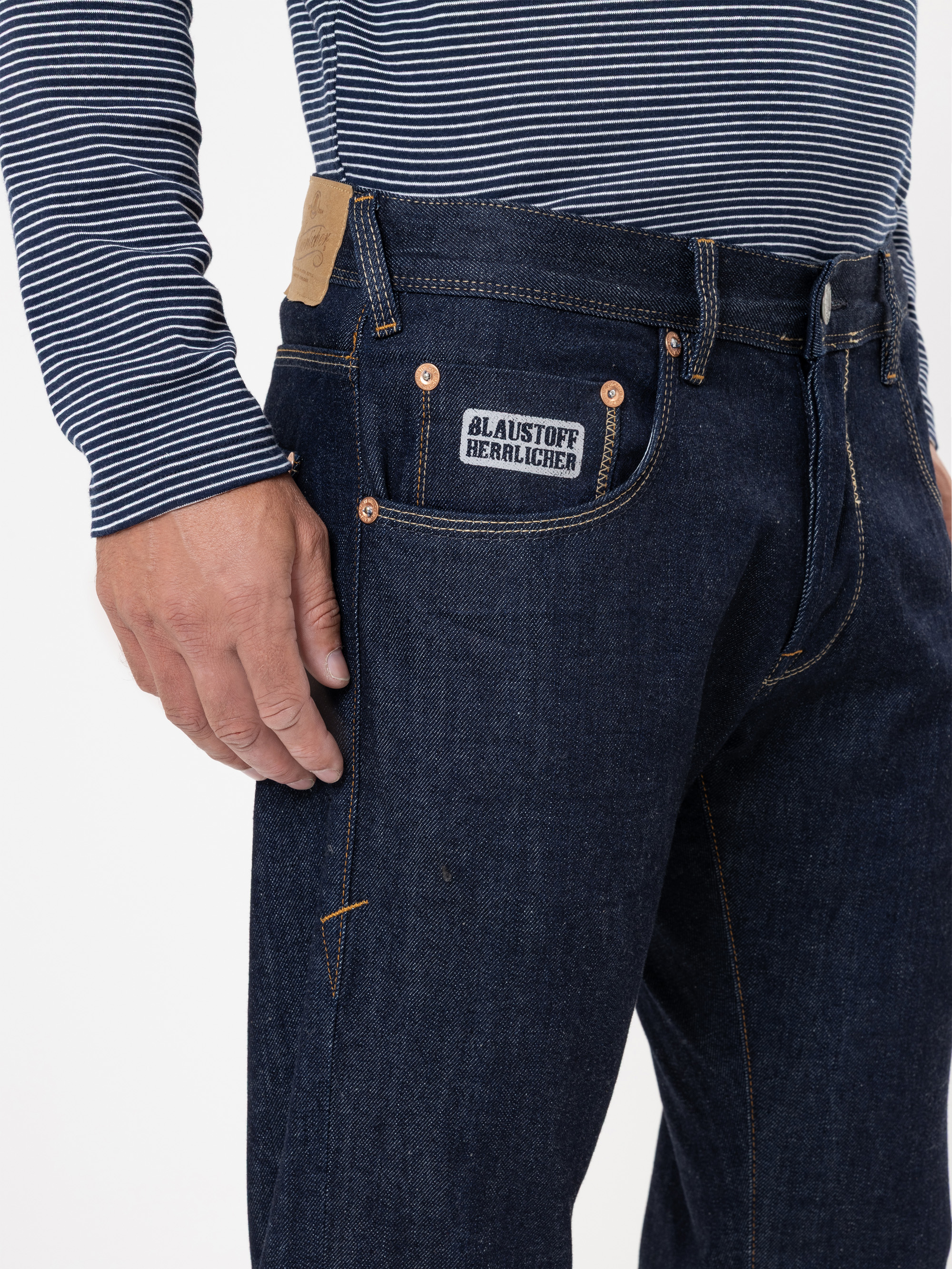 Eine Person, die eine dunkelblaue Jeans mit einem sichtbaren BLAUSTOFF HERLICHER-Etikett auf der Tasche und ein marineblau-weiß gestreiftes langärmeliges Hemd trägt, steht vor einem einfarbigen Hintergrund.