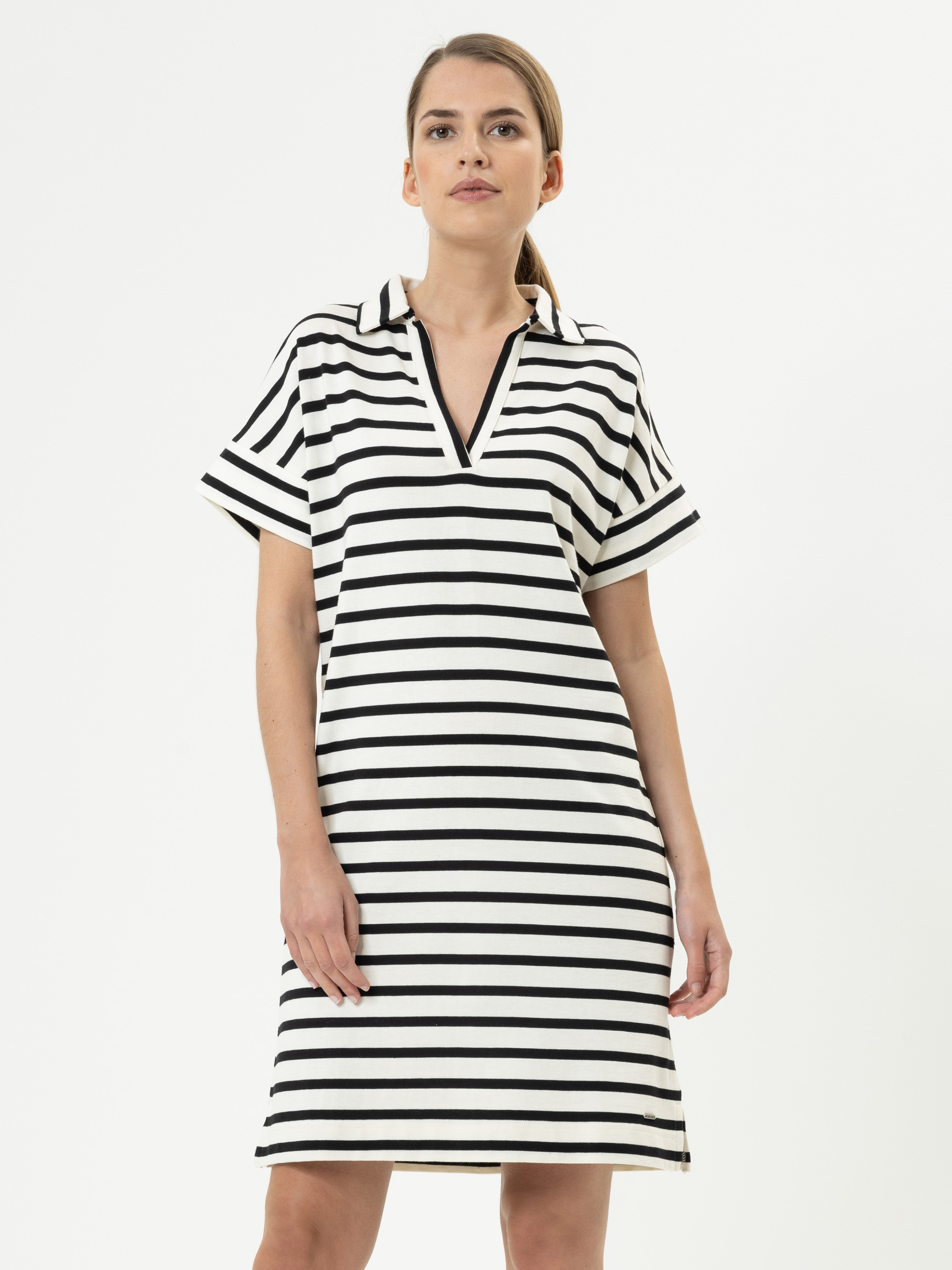 Eine Frau, die ein knielanges, kurzärmeliges, schwarz-weißes, horizontal gestreiftes Kleid mit V-Ausschnitt und Kragen trägt, steht vor einem schlichten weißen Hintergrund.