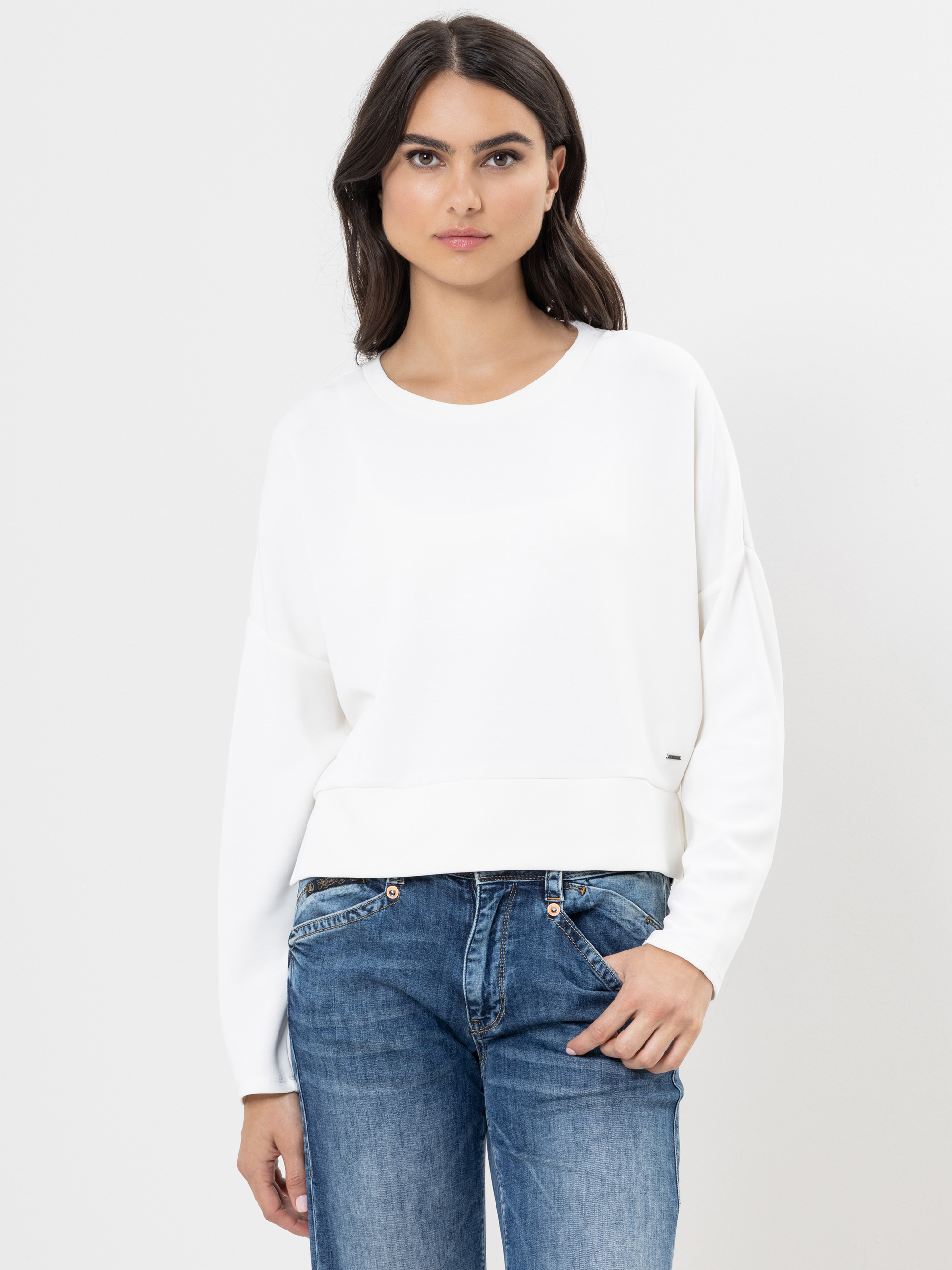 Eine Frau mit langen dunklen Haaren, die ein locker sitzendes weißes Sweatshirt und blaue Jeans trägt, steht vor einem schlichten weißen Hintergrund und schaut mit neutralem Blick direkt in die Kamera.