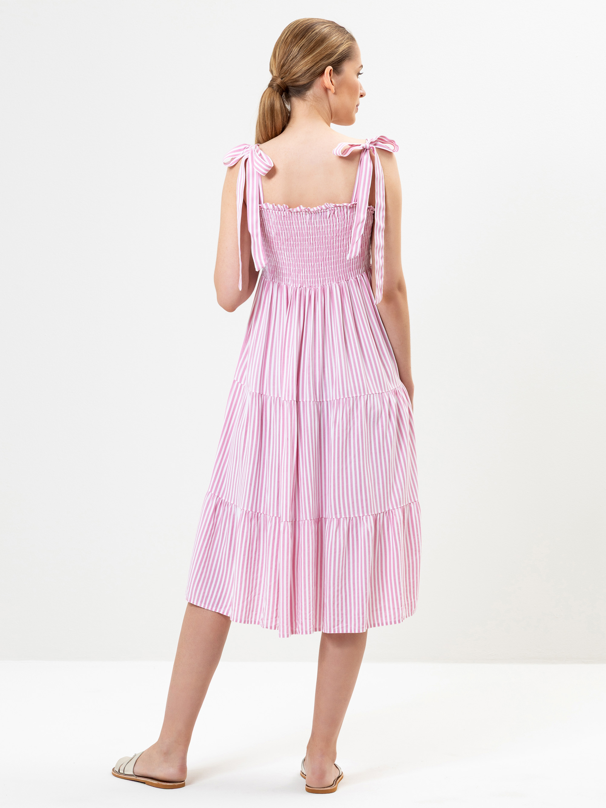 Eine Frau mit hellbraunem, zurückgebundenem Haar trägt ein rosa-weiß gestreiftes, ärmelloses, gestuftes Midikleid mit Schulterriemen. Sie steht mit dem Gesicht zur Wand und trägt weiße flache Sandalen vor einem schlichten weißen Hintergrund.