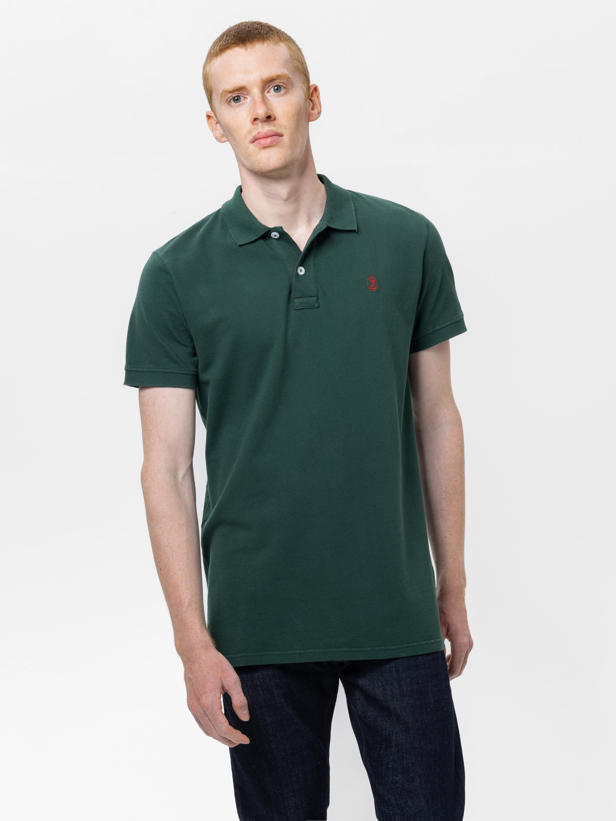 Ein Mann mit kurzen roten Haaren trägt ein dunkelgrünes Polohemd und dunkle Jeans und steht mit neutralem Gesichtsausdruck vor einem schlichten weißen Hintergrund.