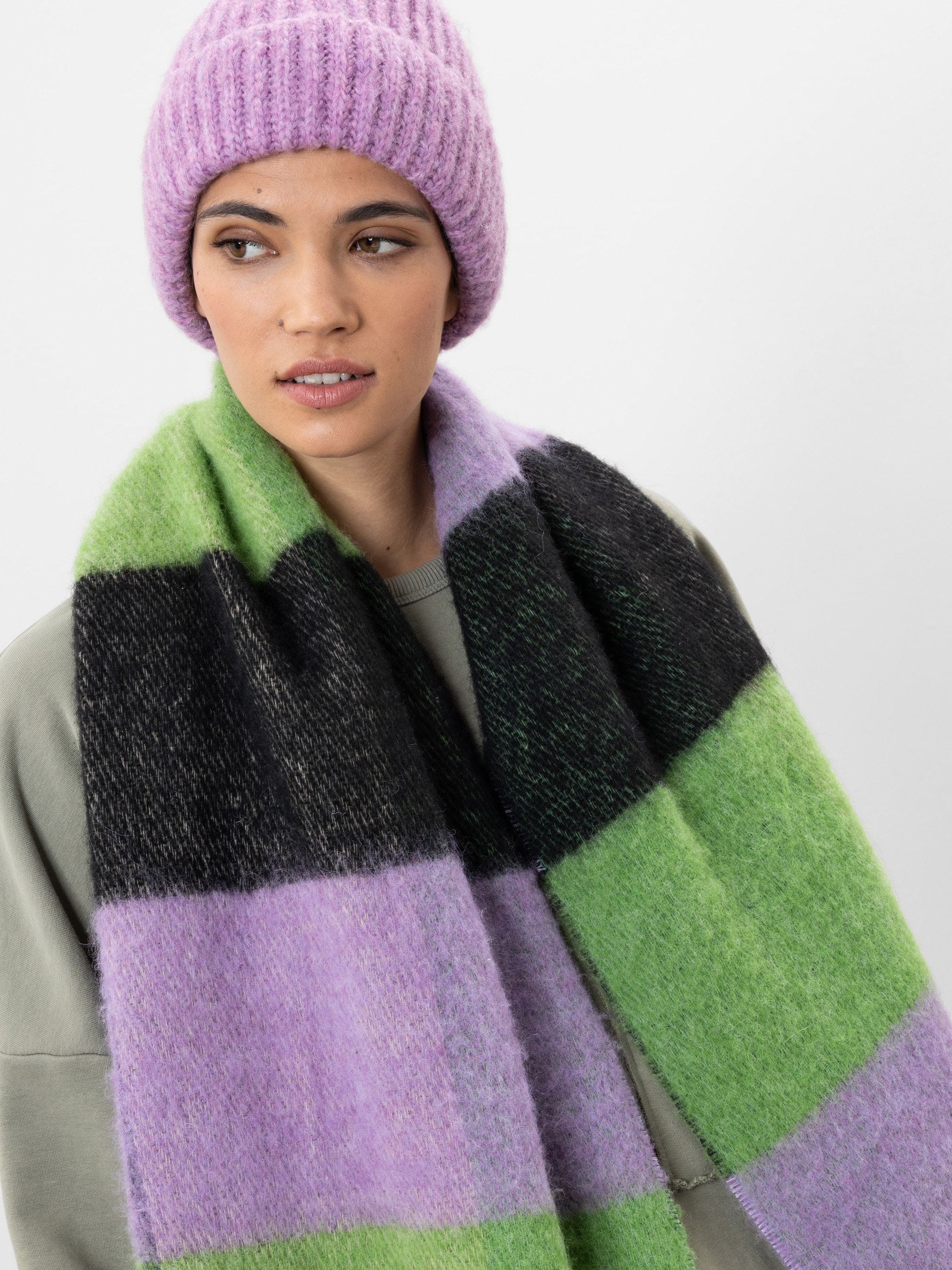 Eine Frau, die eine lilafarbene Strickmütze, einen grauen Pullover und einen wuscheligen Schal mit großen grünen, schwarzen und lilafarbenen Blöcken trägt, steht vor einem schlichten weißen Hintergrund und schaut leicht nach links.